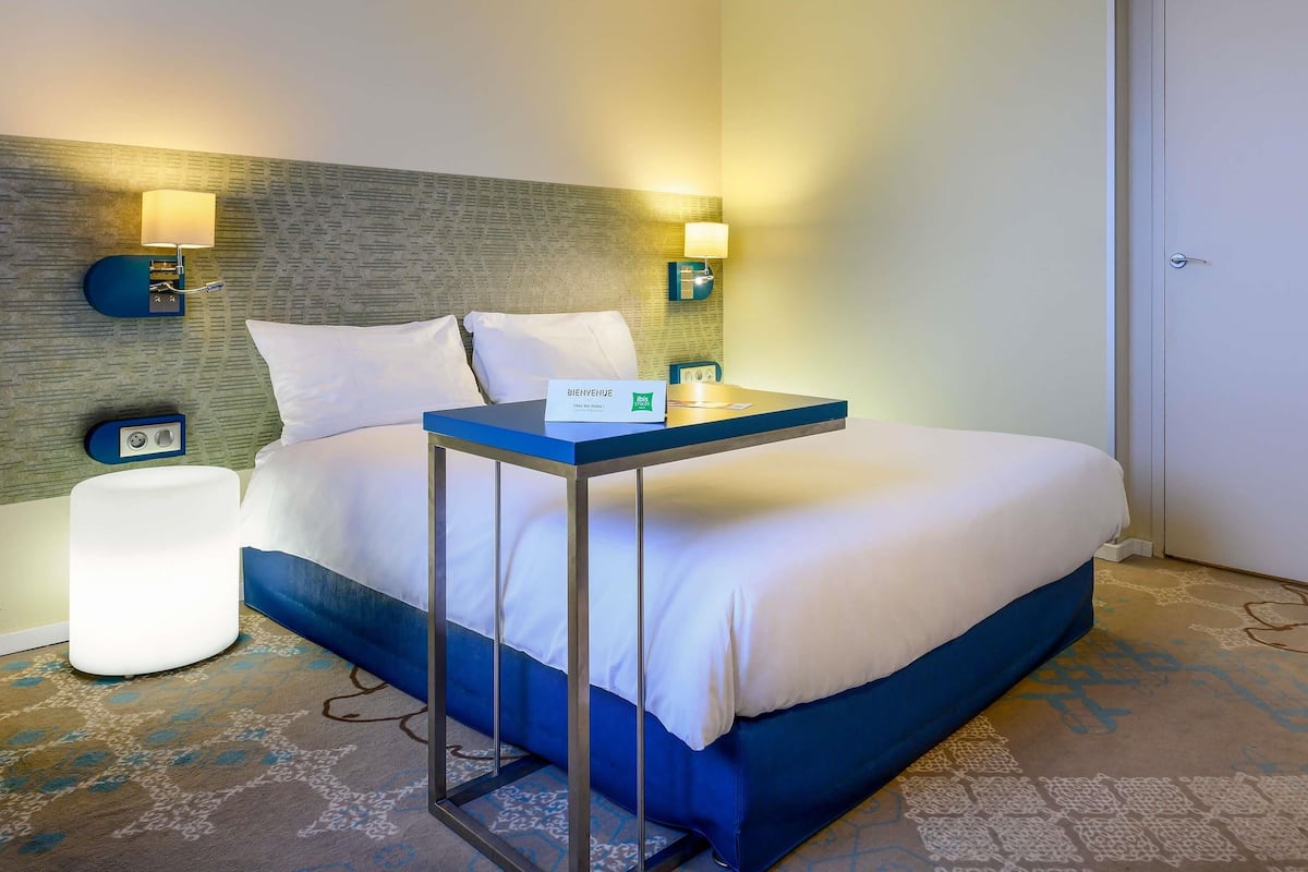 A hotel room with a bed, bedside table, lamp, and a welcome sign.