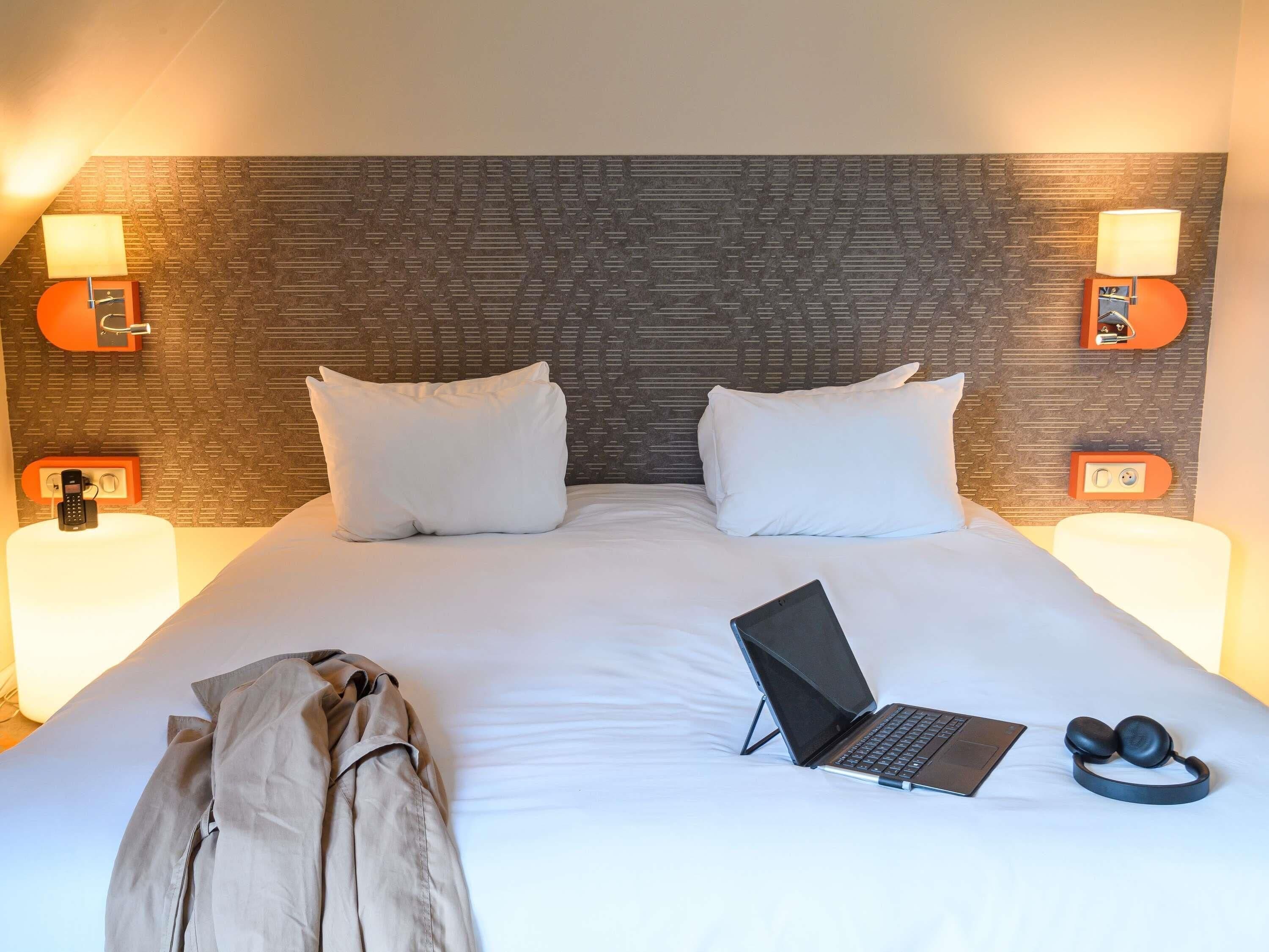 Ein Hotelzimmer mit einem Bett, zwei Nachttischlampen, einem Laptop, Kopfhörern und einem Koffer.