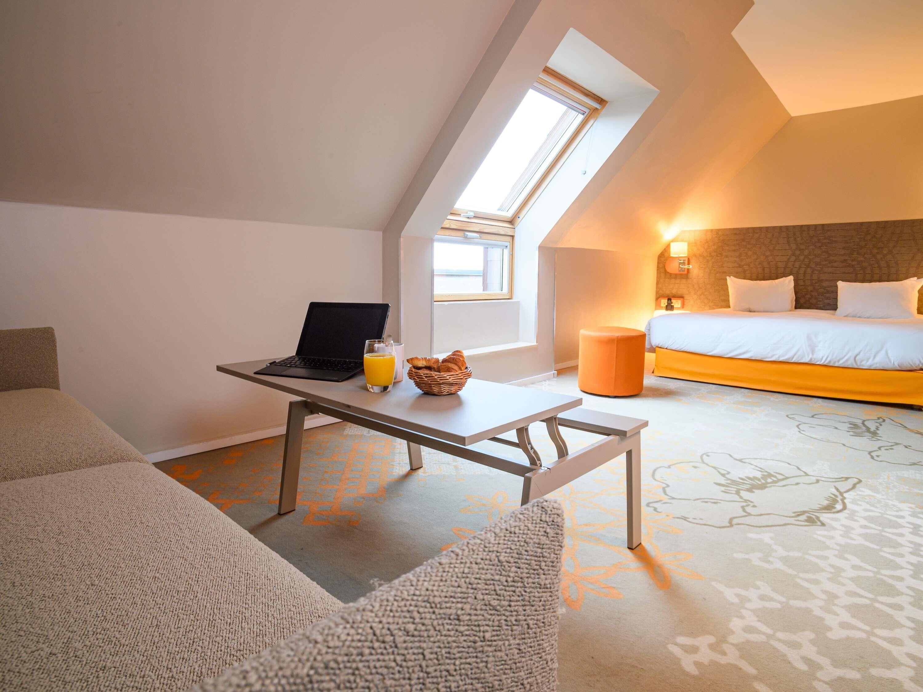 Una habitación de hotel moderna con una cama tipo loft, un ordenador portátil sobre una mesita pequeña, un vaso de zumo de naranja y una cesta de mimbre.