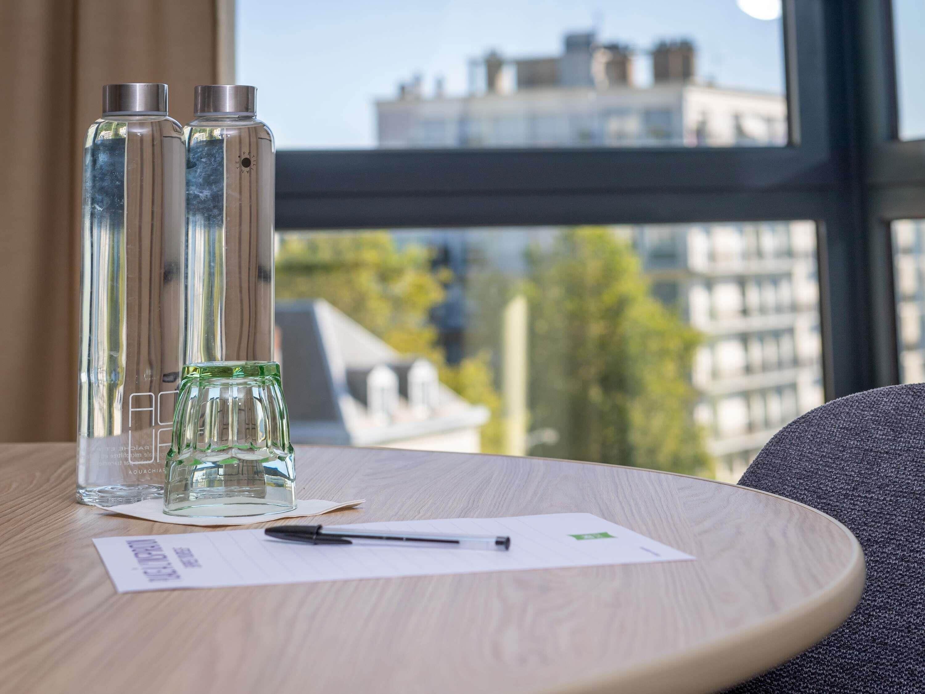 A table with two clear bottles, a pen, and a piece of paper in a room with a view of buildings through a window.