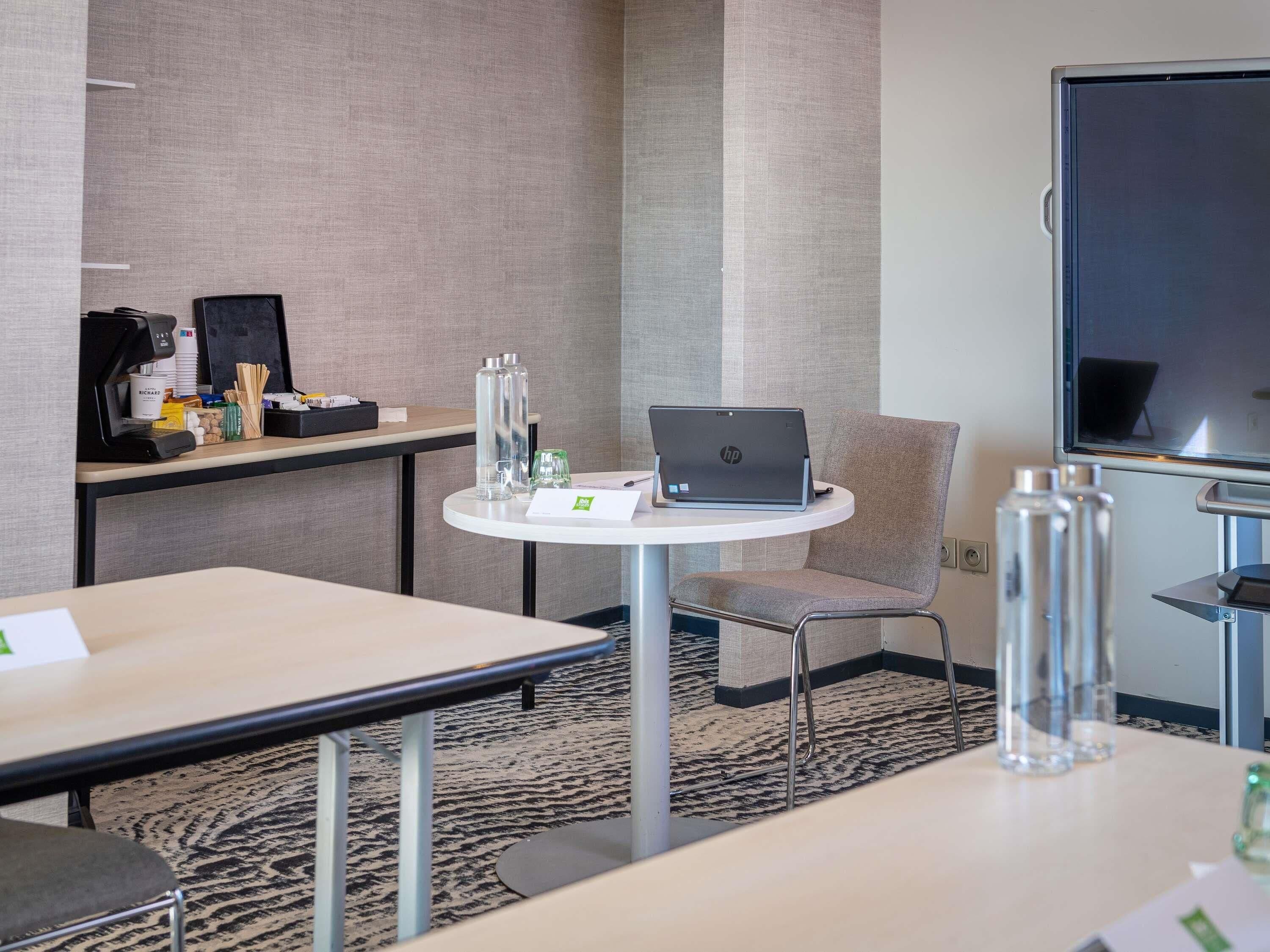 A hotel room with a round table, a laptop, a chair, a coffee machine, a projector screen, and a carpeted floor.