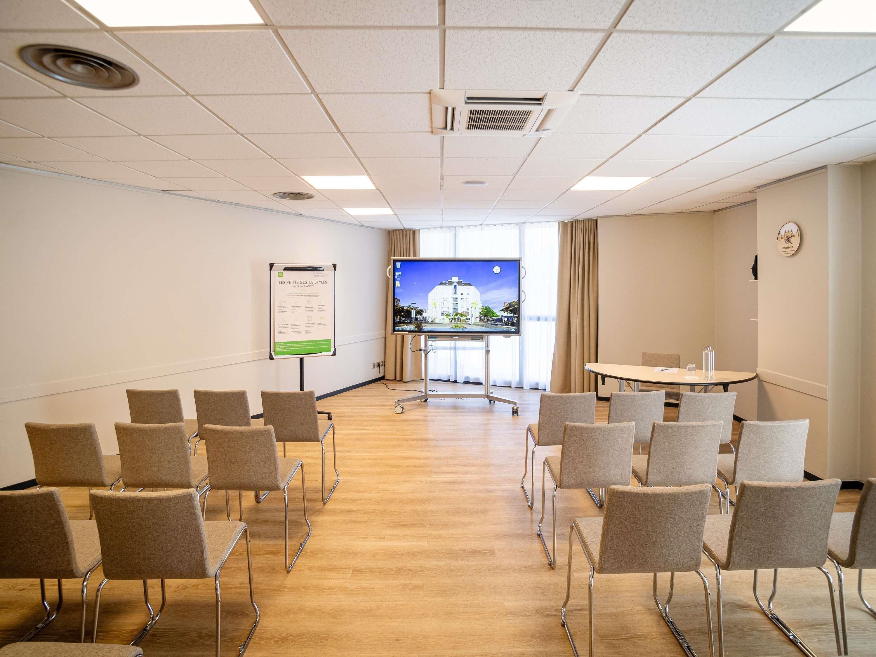 Une salle de conférence équipée d’un grand écran, d’un tableau blanc et de nombreuses chaises disposées pour une réunion.