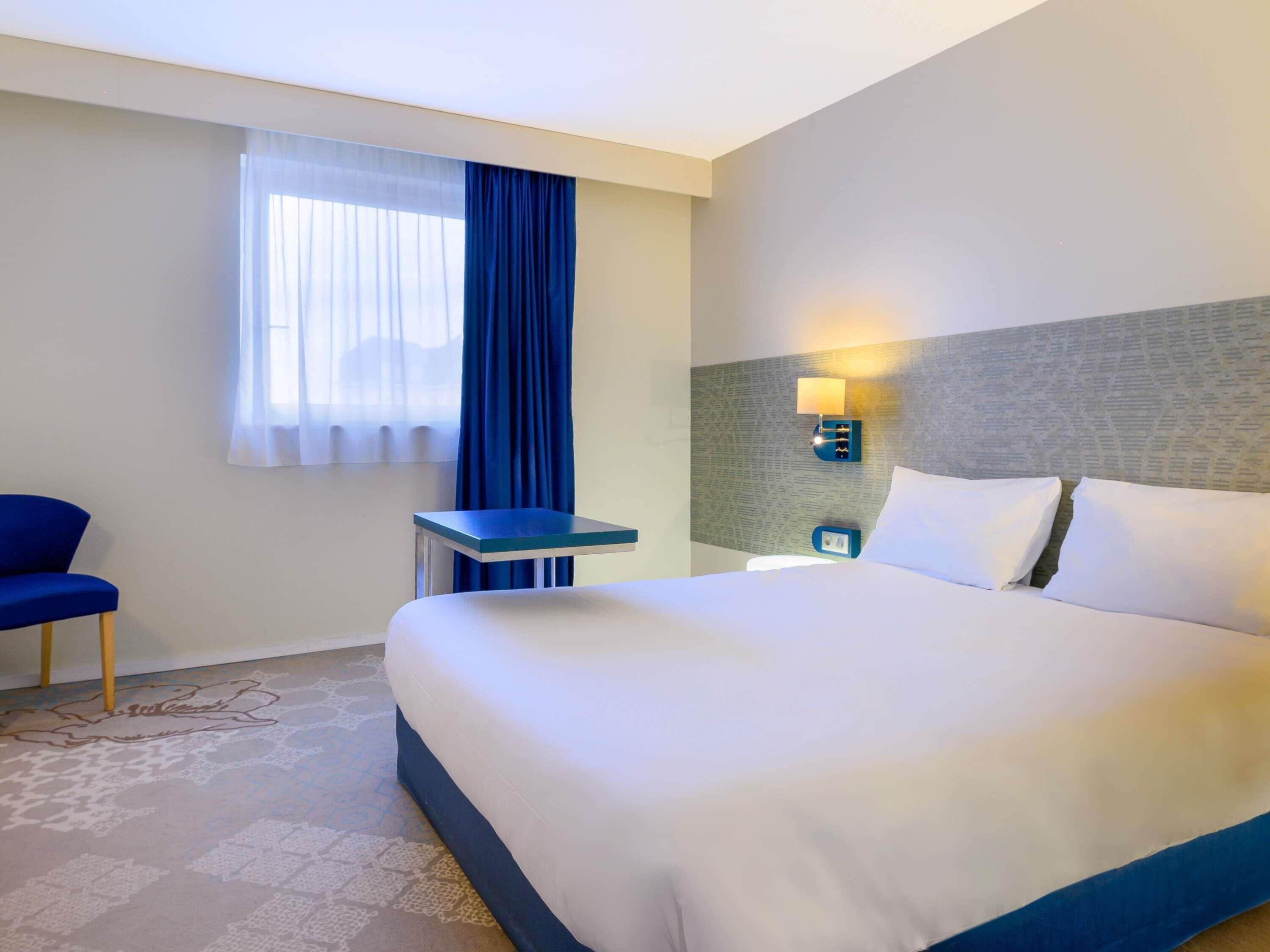 Quarto de hotel com cama, escrivaninha, cadeira, janela com cortinas e luminária fixada na parede.