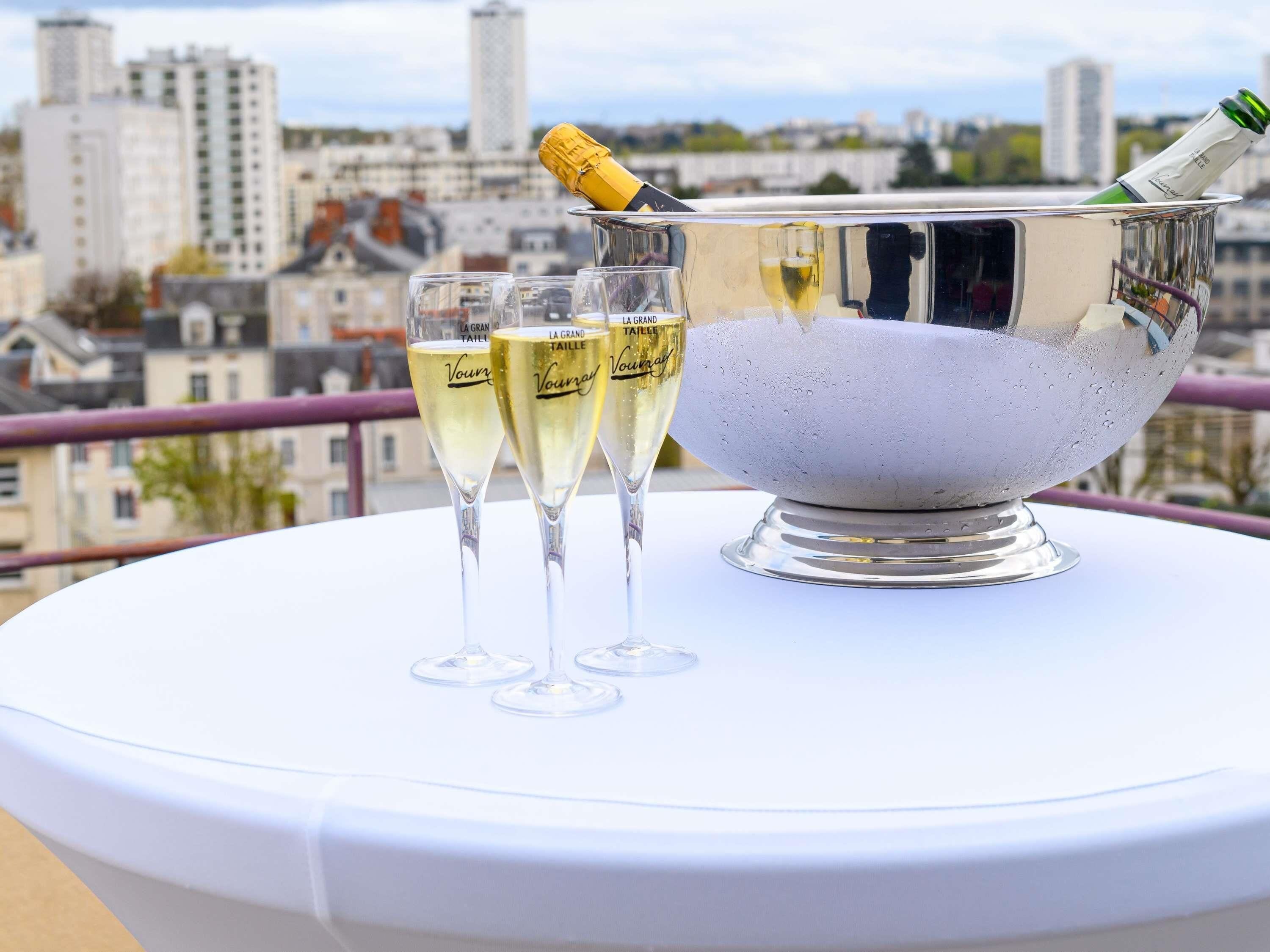 Ein Tisch mit einem silbernen Champagnerkühler und drei Champagnergläsern auf einer weißen Tischdecke, mit Blick auf eine Stadtlandschaft.