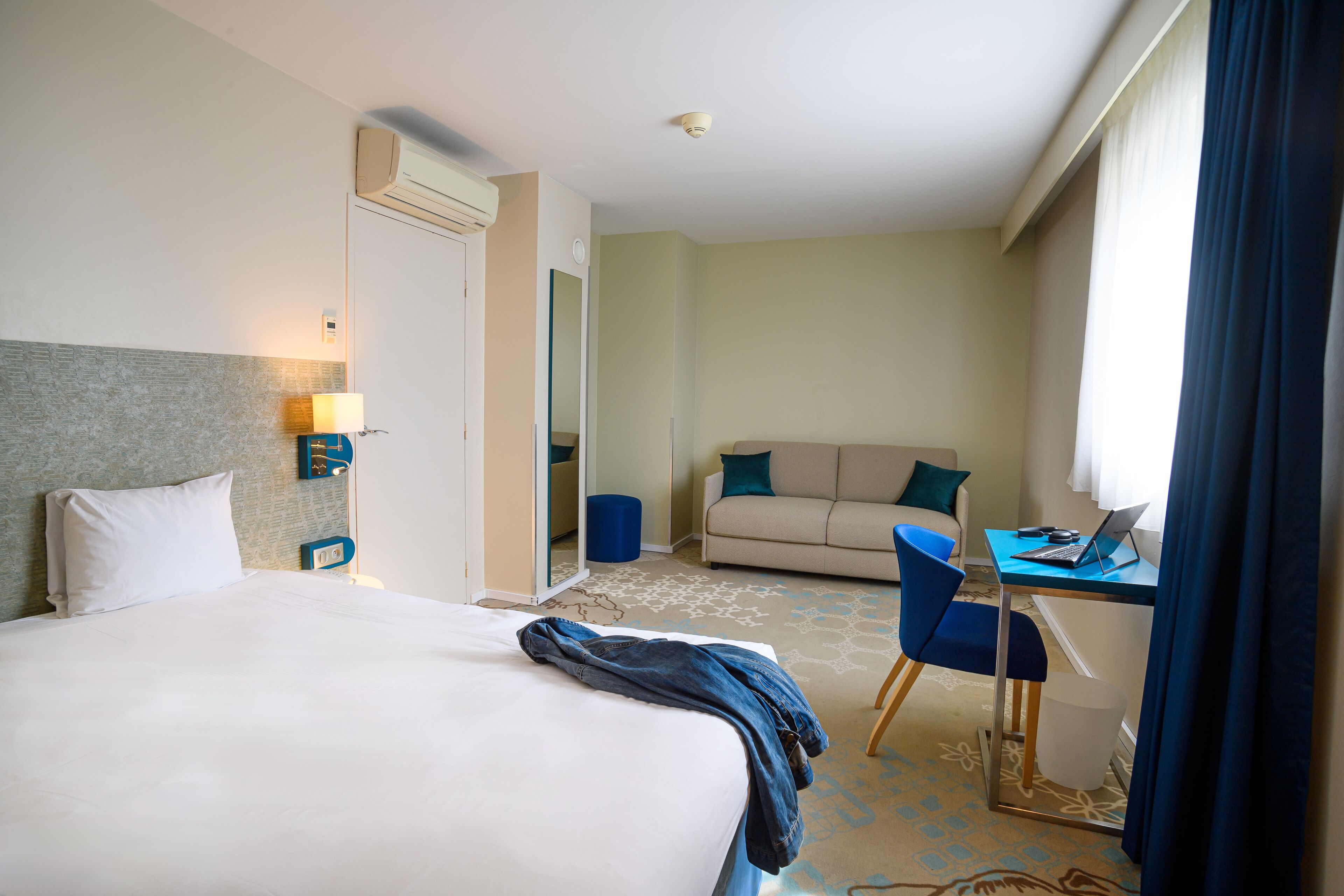 A hotel room with a bed, a desk with a laptop, a sofa, and a mirror.