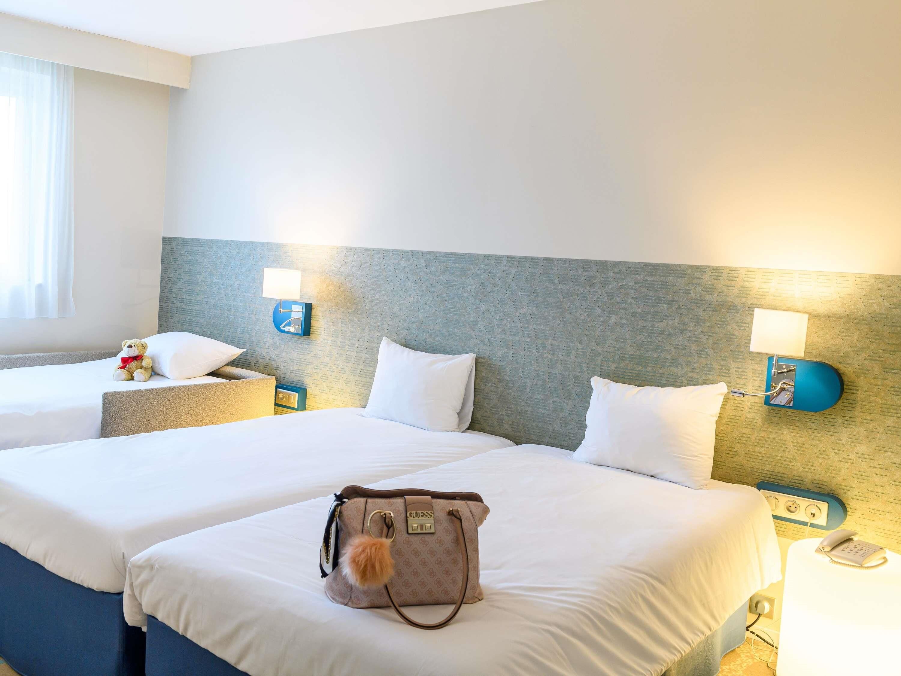 Una camera d'albergo con due letti, un comodino, una lampada, un orsetto di peluche e una borsa.