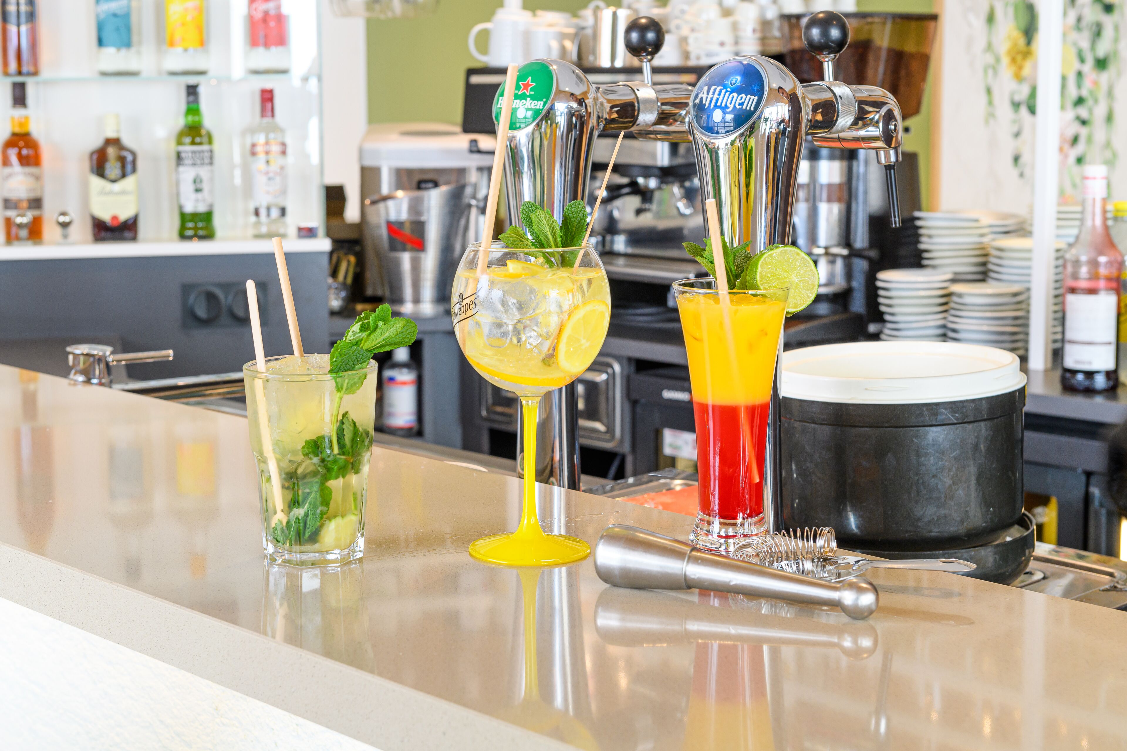 Una zona de bar con tres grifos sirviendo diferentes bebidas, un coctelero y una pila de platos al fondo.