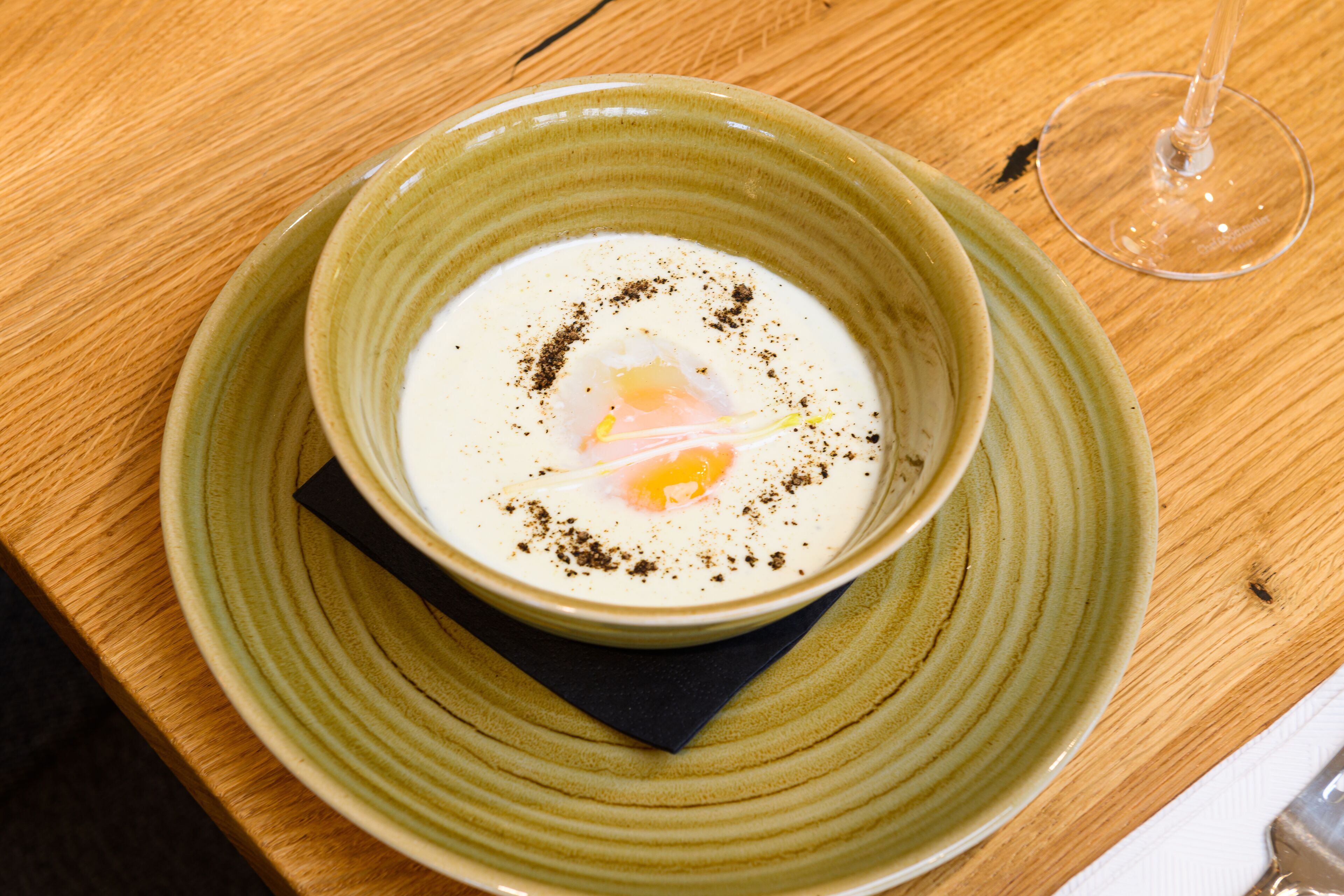 A bowl of soup with a poached egg on a green plate, accompanied by a wine glass and a napkin on a wooden table.
