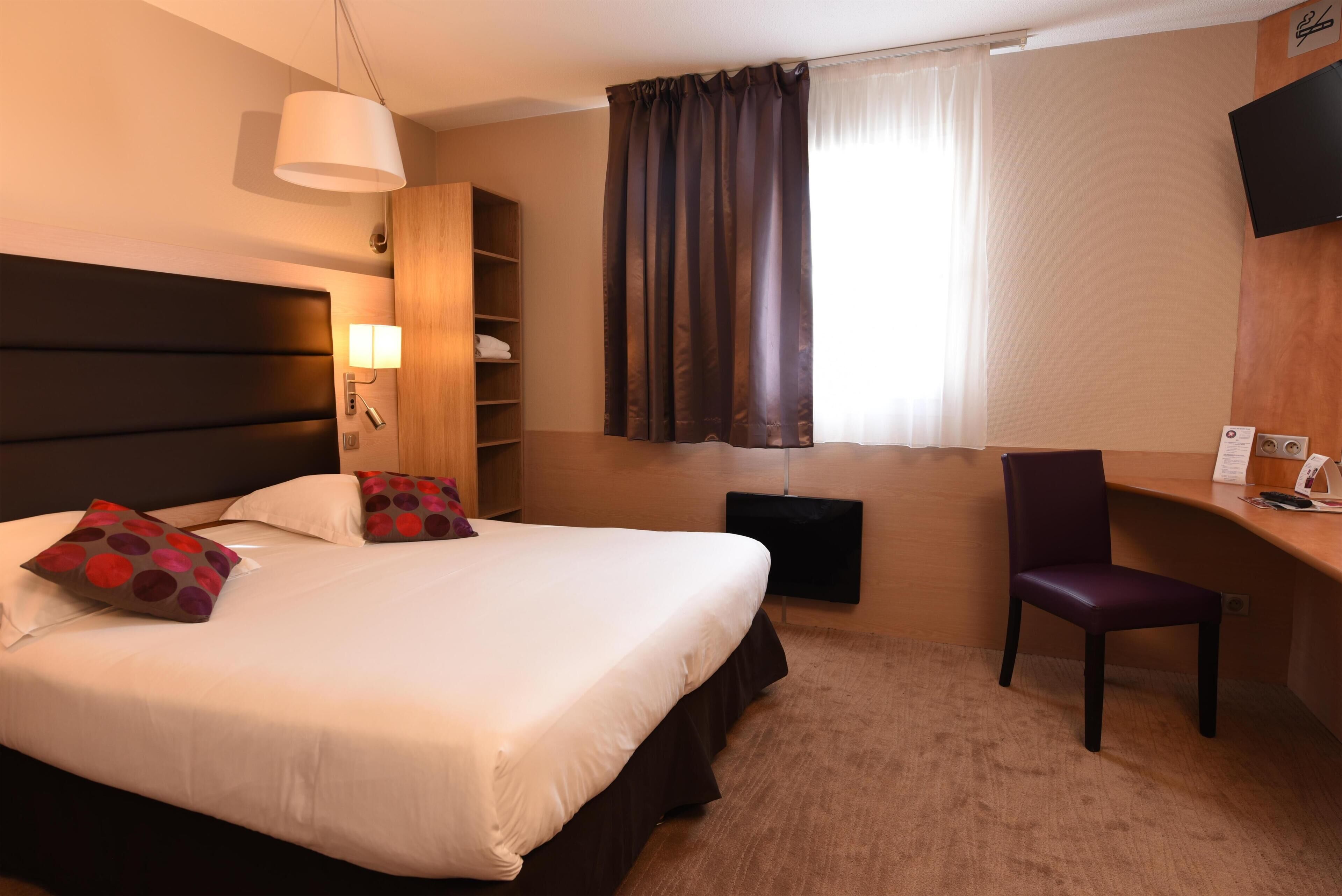 A hotel room with a large bed, a desk, a chair, a TV, a bookshelf, and a window with curtains.