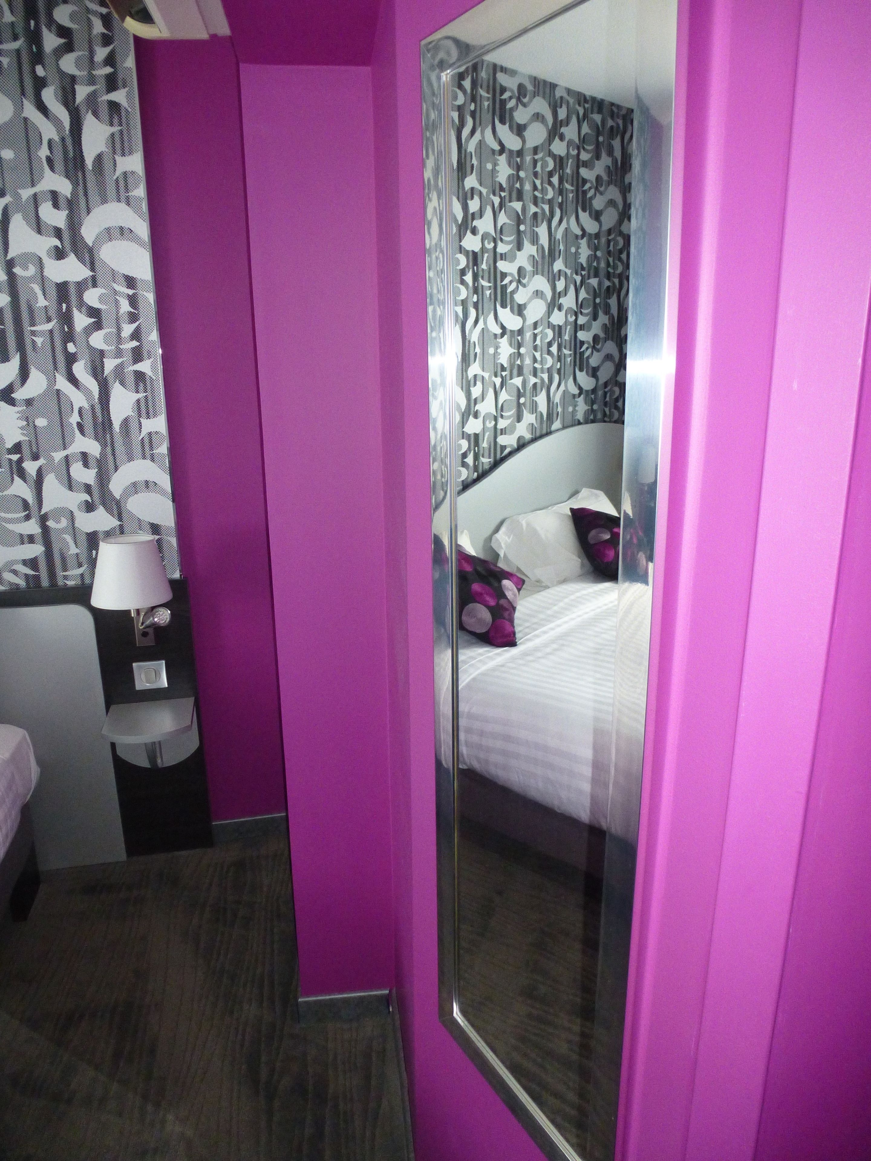 A hotel room with a large mirror, a bed with white bedding and colorful pillows, a bedside table with a lamp, and a patterned curtain.
