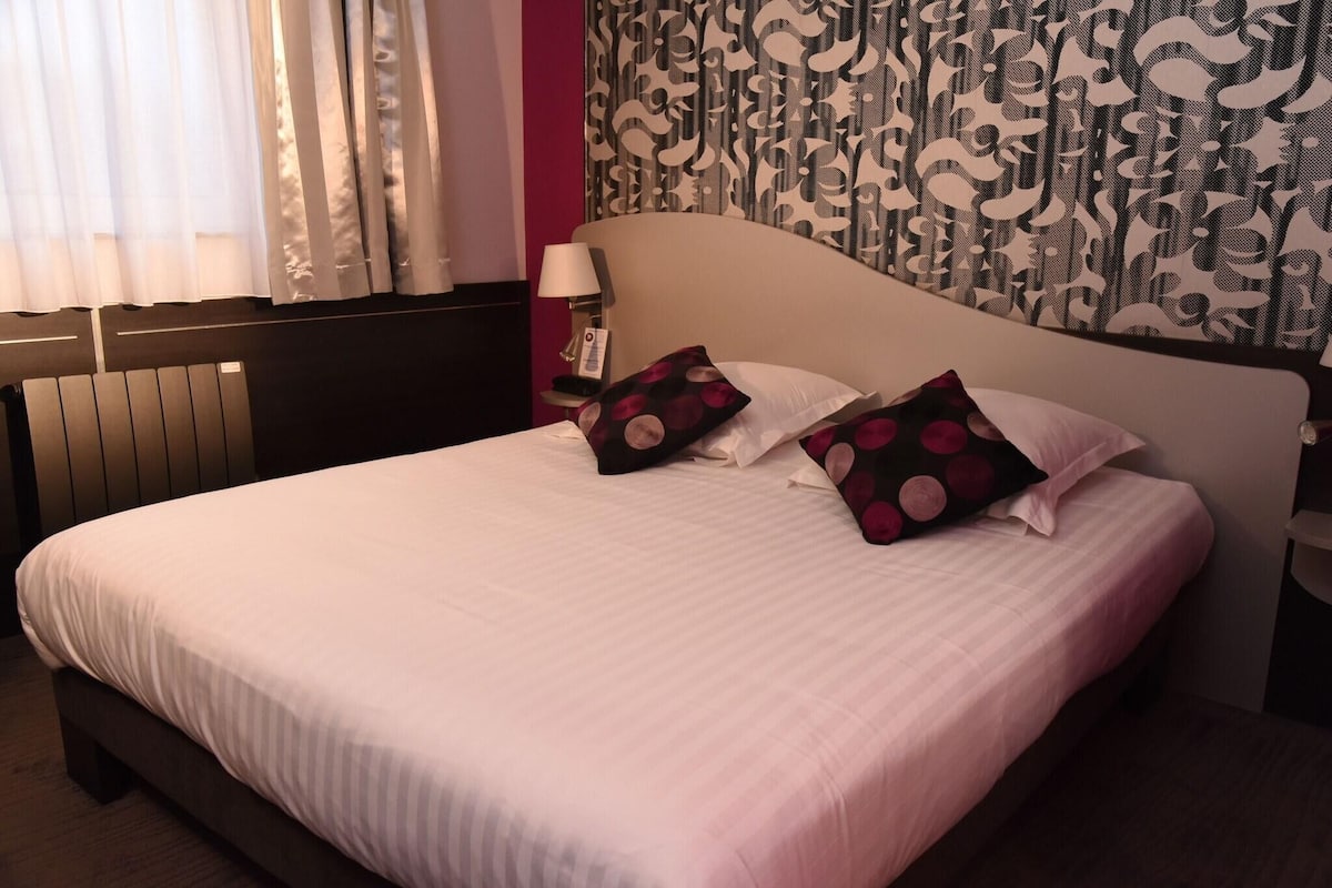 Una cama bien hecha con ropa blanca, cabecera con diseño estampado y mesita de noche con lámpara y teléfono.