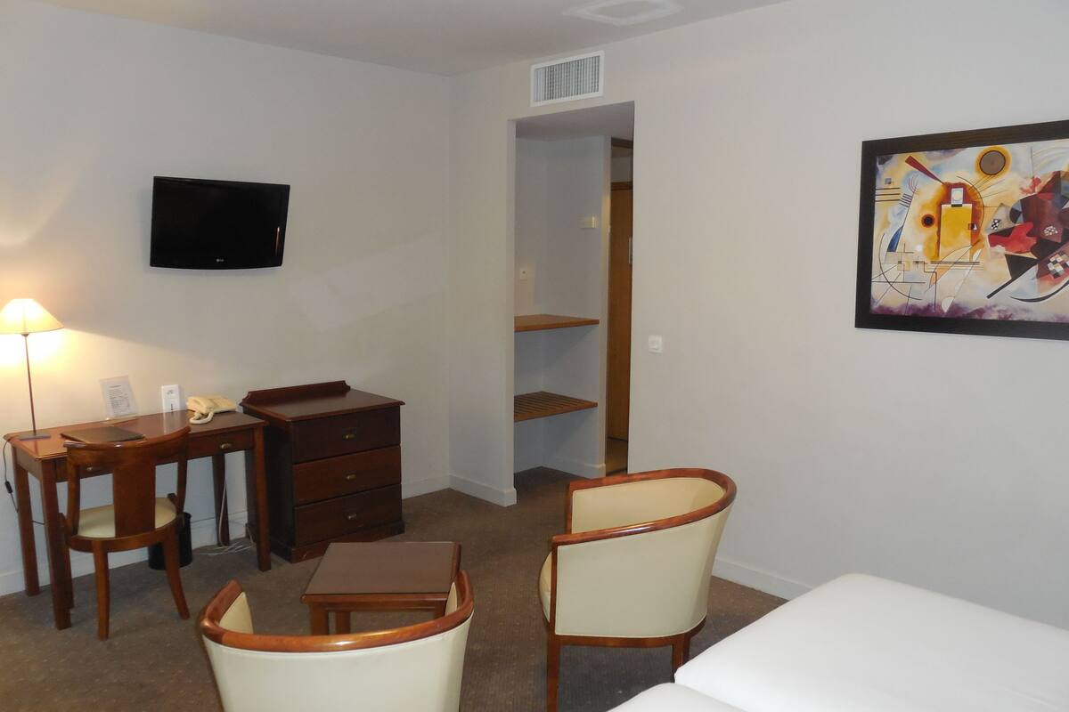 A hotel room with a bed, a desk with a chair, a small table, a lamp, a TV, and a painting on the wall.