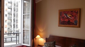 A hotel room with a bed, a window with a view of buildings, a lamp, and a painting on the wall. - Hôtel Cardinal Rive Gauche (Paris)