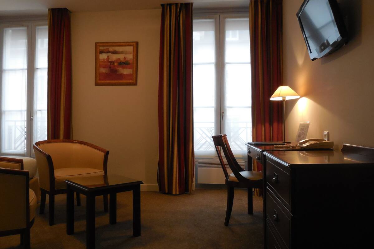 A hotel room with a desk, two chairs, a television mounted on the wall, and a window with curtains.