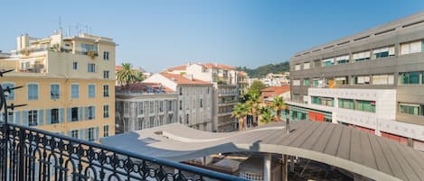 A balcony with wrought iron railings overlooking a cityscape with various buildings, including a modern structure with a flat roof and a traditional building with a sloped roof.