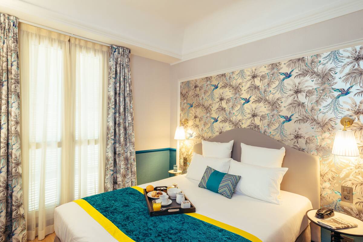 A hotel room with a bed, a tray of breakfast items, a nightstand with a lamp, and a floral wallpaper.