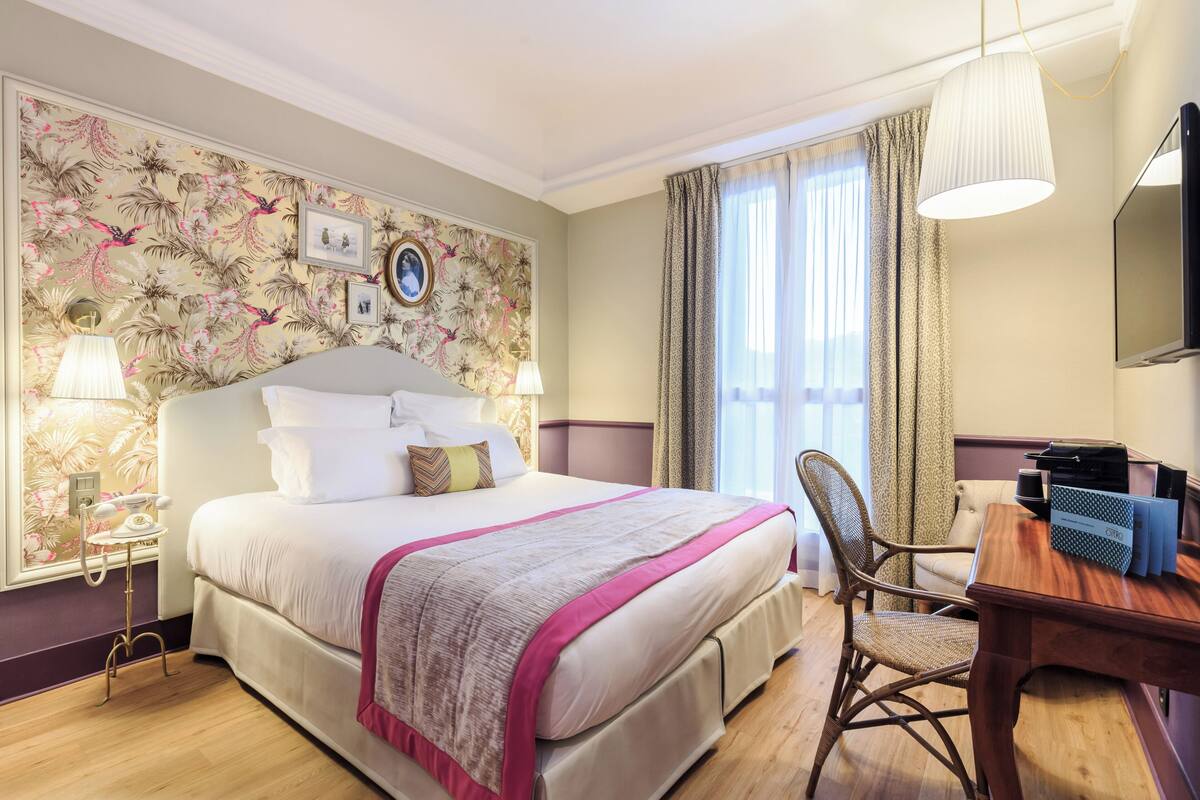 A hotel room with a large bed, a desk, a chair, a television, and a window with curtains.