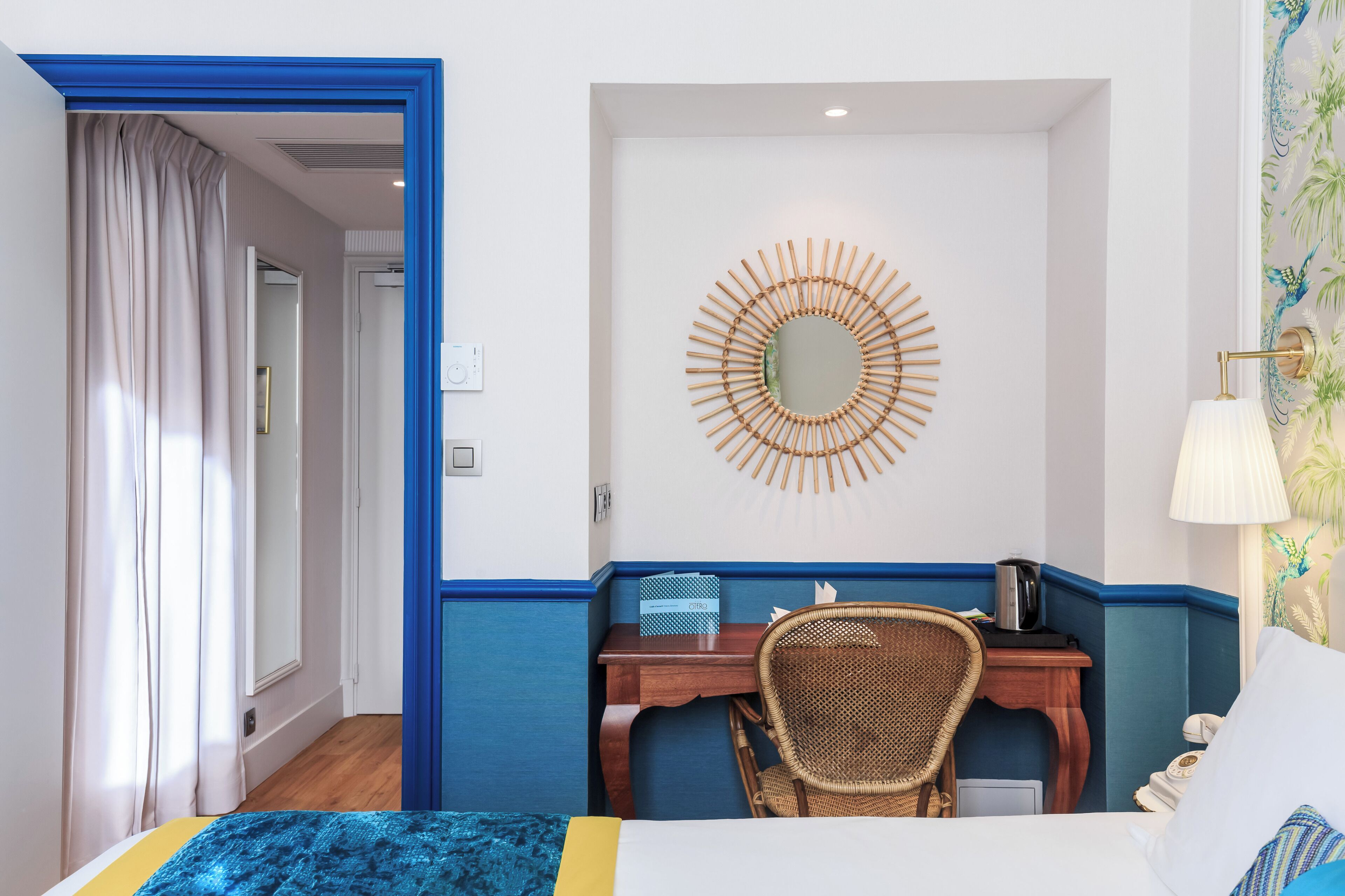 A hotel room with a bed, a desk with a chair, a wall-mounted mirror, and a lamp.