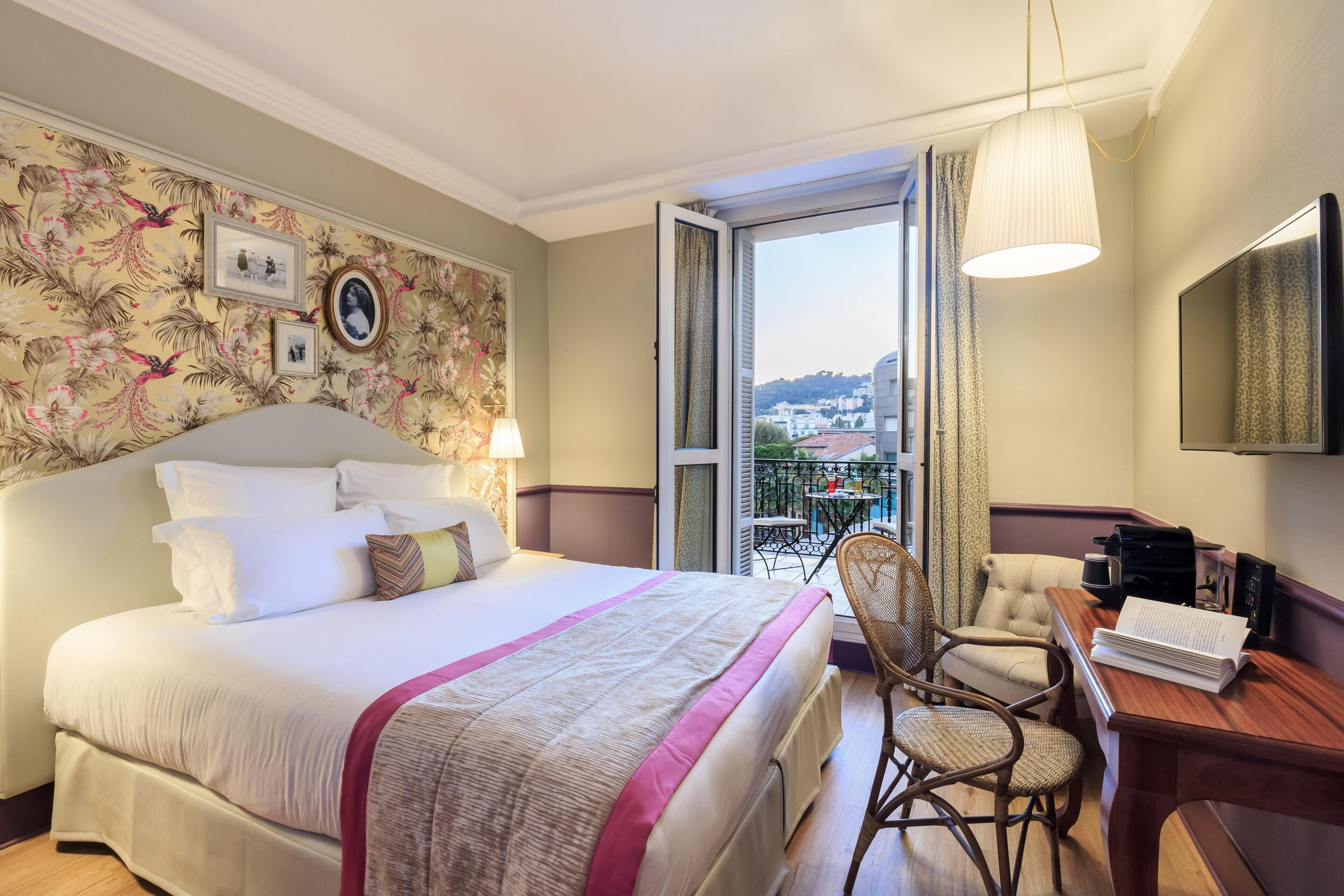 A hotel room with a large bed, a desk with a chair, a balcony with a view of buildings, and a mounted TV.