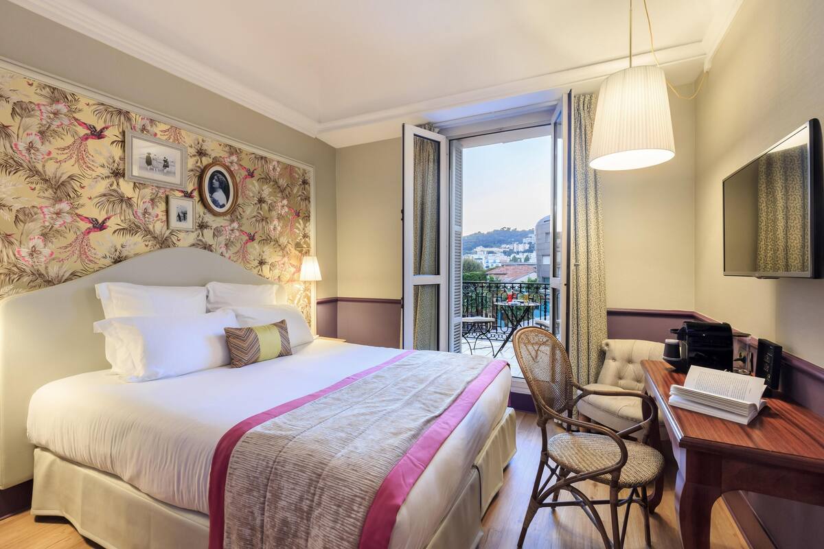 A hotel room with a large bed, a desk with a chair, a balcony with a view of buildings, and a mounted TV.
