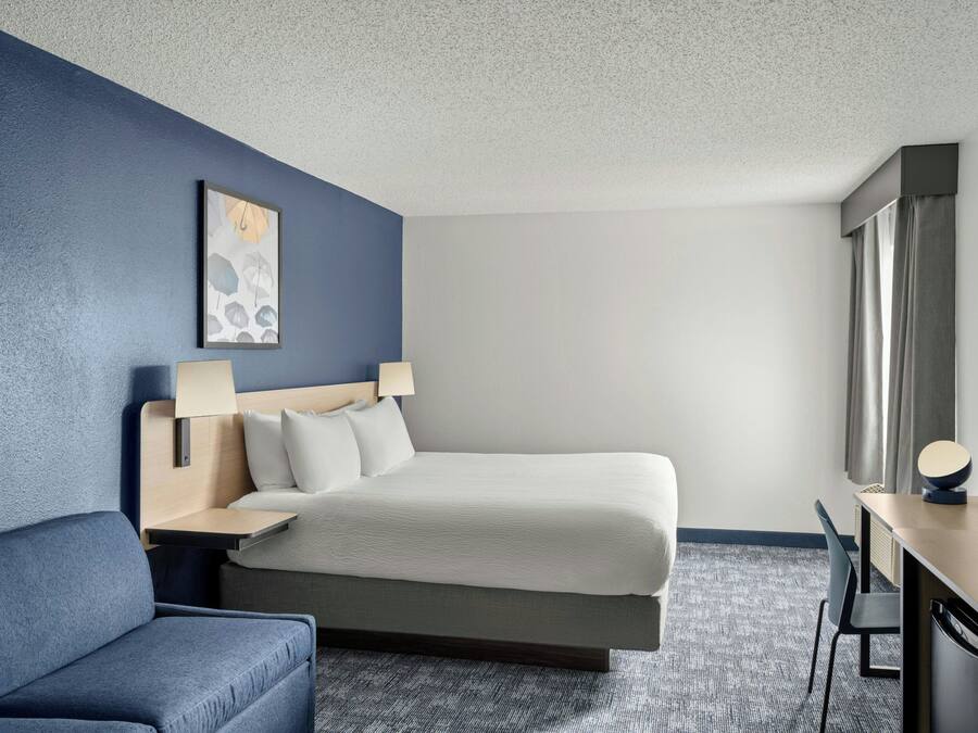 A hotel room with a bed, a desk, a chair, a TV, and a painting on the wall.