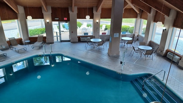 Una piscina interior con sillones y mesas, vigas de madera en el techo y grandes ventanales con vistas a un estacionamiento.