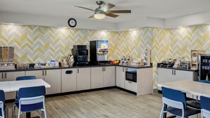 Una cocina moderna con un revestimiento de pared en patrón de espiga, ventilador de techo, reloj y varios electrodomésticos, incluyendo una cafetera y un microondas.