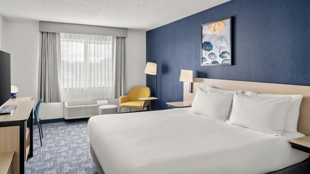 A hotel room with a large bed, a desk, a chair, a bedside table, a lamp, and a window with curtains. - City Express By Marriott Freeport (Freeport)