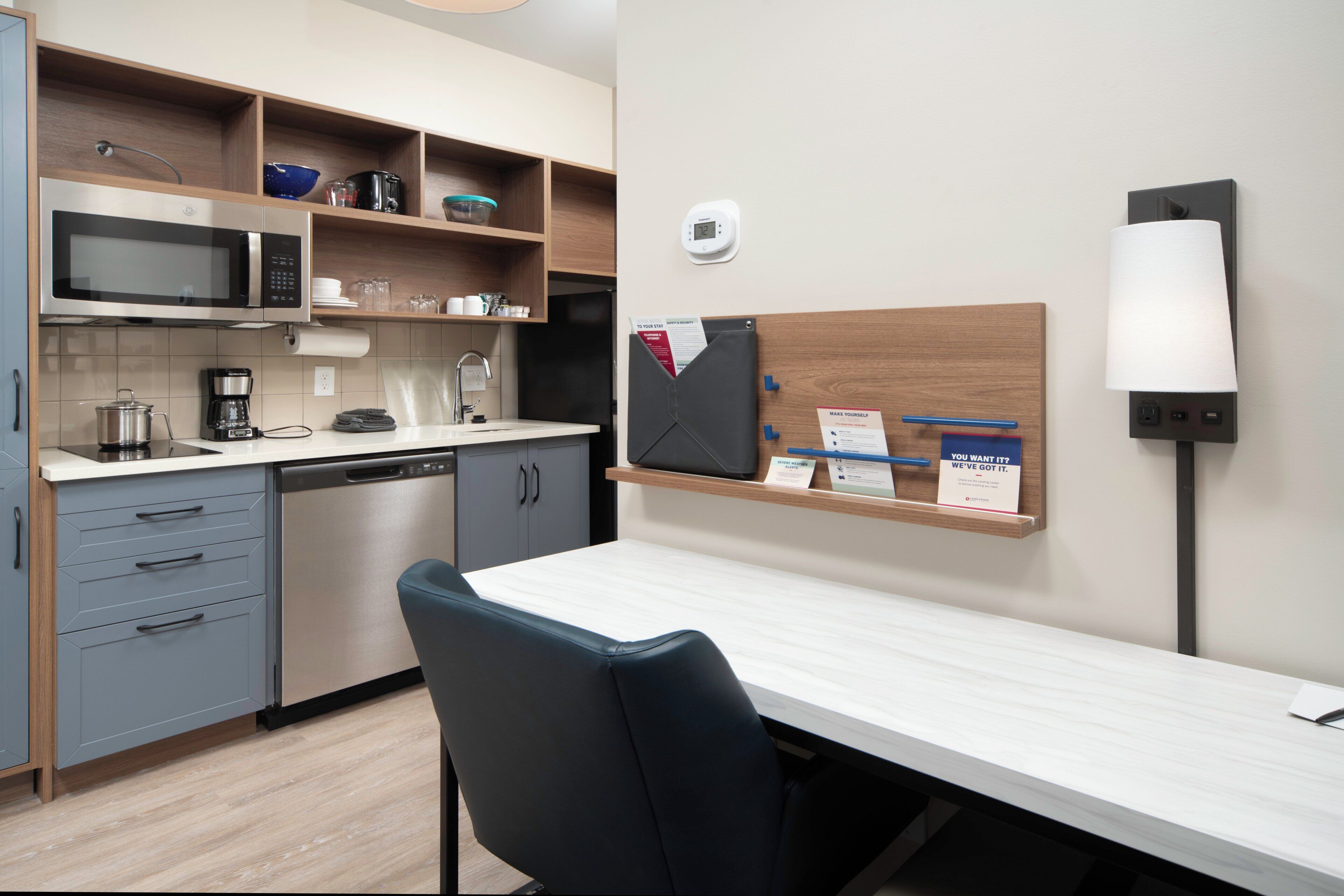 A modern kitchen with a microwave, coffee maker, and a small refrigerator. There is a wooden shelf above the counter with various items.