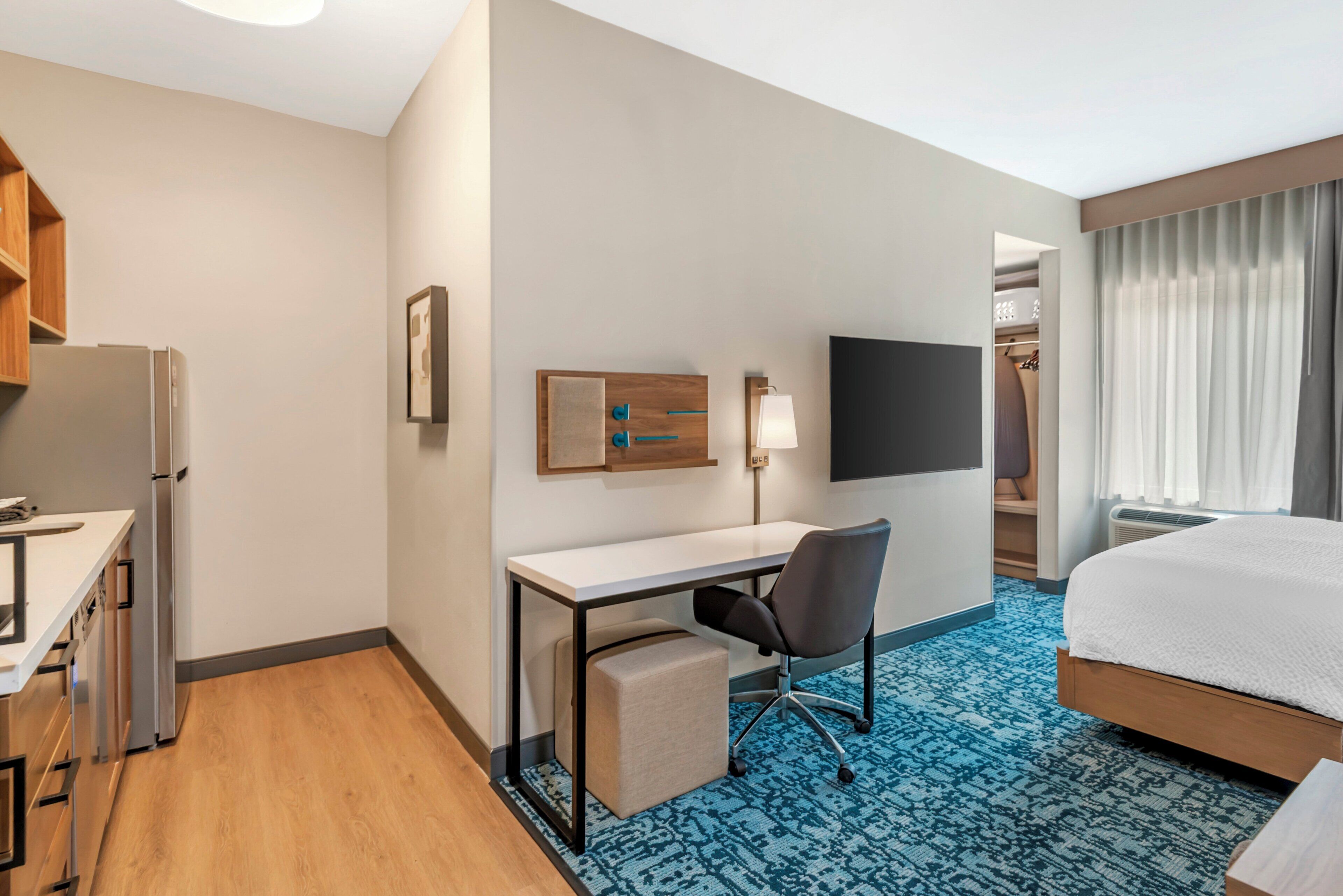 A hotel room with a bed, a desk, a chair, a flat-screen TV, a wardrobe, and a small kitchenette.