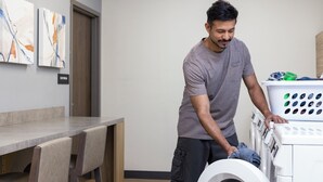 A person loading clothes into a front-loading washing machine in a room with a table and abstract art on the wall. - Candlewood Suites Rowlett - Lake Ray Hubbard by IHG (Rowlett)