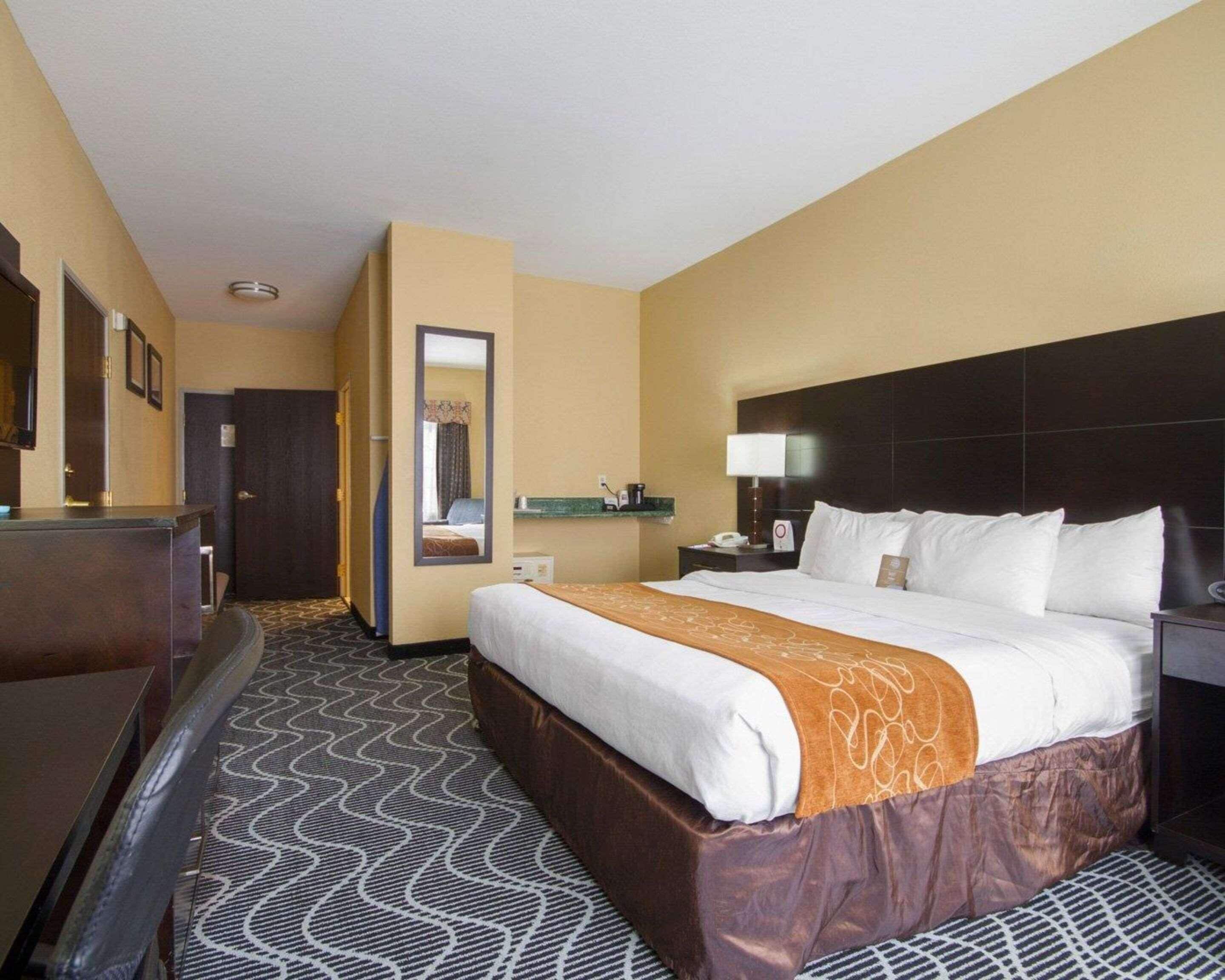 A hotel room with a large bed, a desk, a mirror, and a nightstand with a lamp.