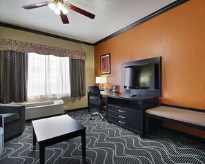 A hotel room with a ceiling fan, a television, a desk, a chair, a sofa, and patterned carpet.