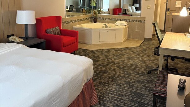 A hotel room with a large bed, a red armchair, a small desk, a mirror, a bathroom with a bathtub, and a shower. - Country Inn & Suites by Radisson, Elgin, IL (Elgin)