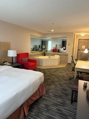 A hotel room with a large bed, a red armchair, a small desk, a mirror, a bathroom with a bathtub, and a shower.