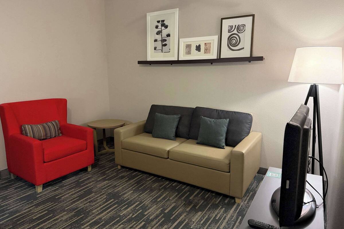 A living room with a red armchair, a beige sofa with cushions, a small round table, a flat-screen TV, and a wall-mounted shelf with framed pictures.