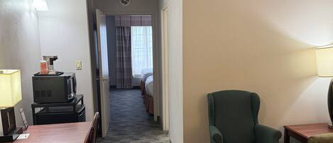 A hotel room with a dining area, a green armchair, a TV, and a desk with a lamp.