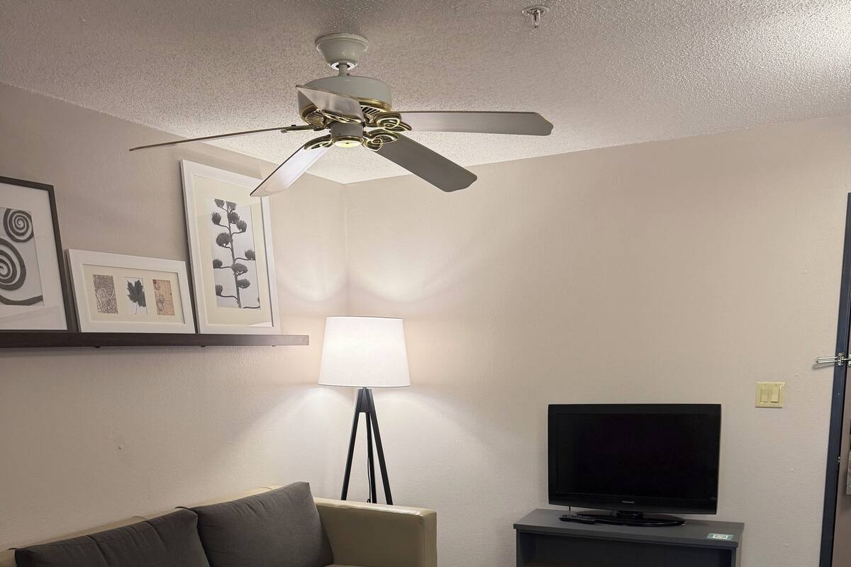 A hotel room with a beige sofa, a red armchair, a standing lamp, a flat-screen TV, and a ceiling fan.