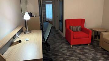 Habitación de hotel con un escritorio de madera, un sillón rojo, una mesita redonda y un banco a lo largo de un pasillo.