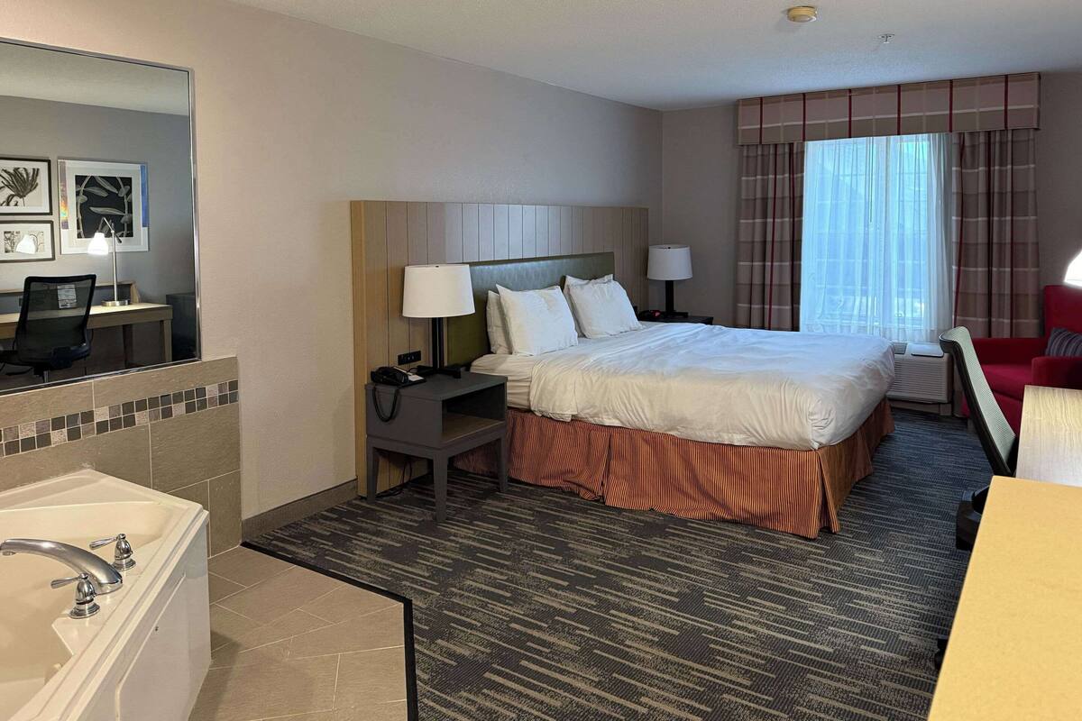 A hotel room with a large bed, a desk with a chair, a desk lamp, a telephone, and a bathroom with a bathtub.