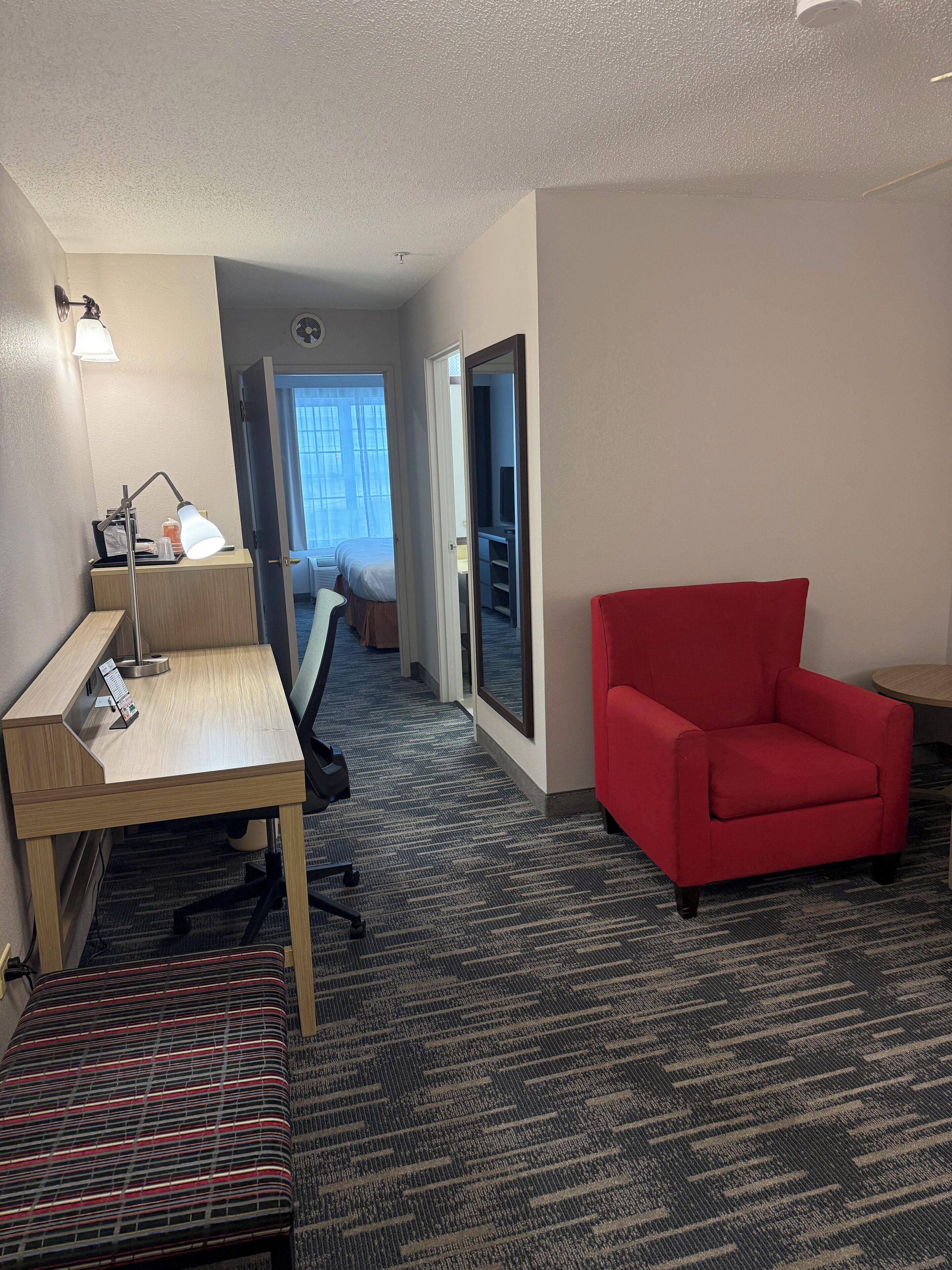 A hotel room with a wooden desk, a red armchair, a standing lamp, and a carpeted floor.