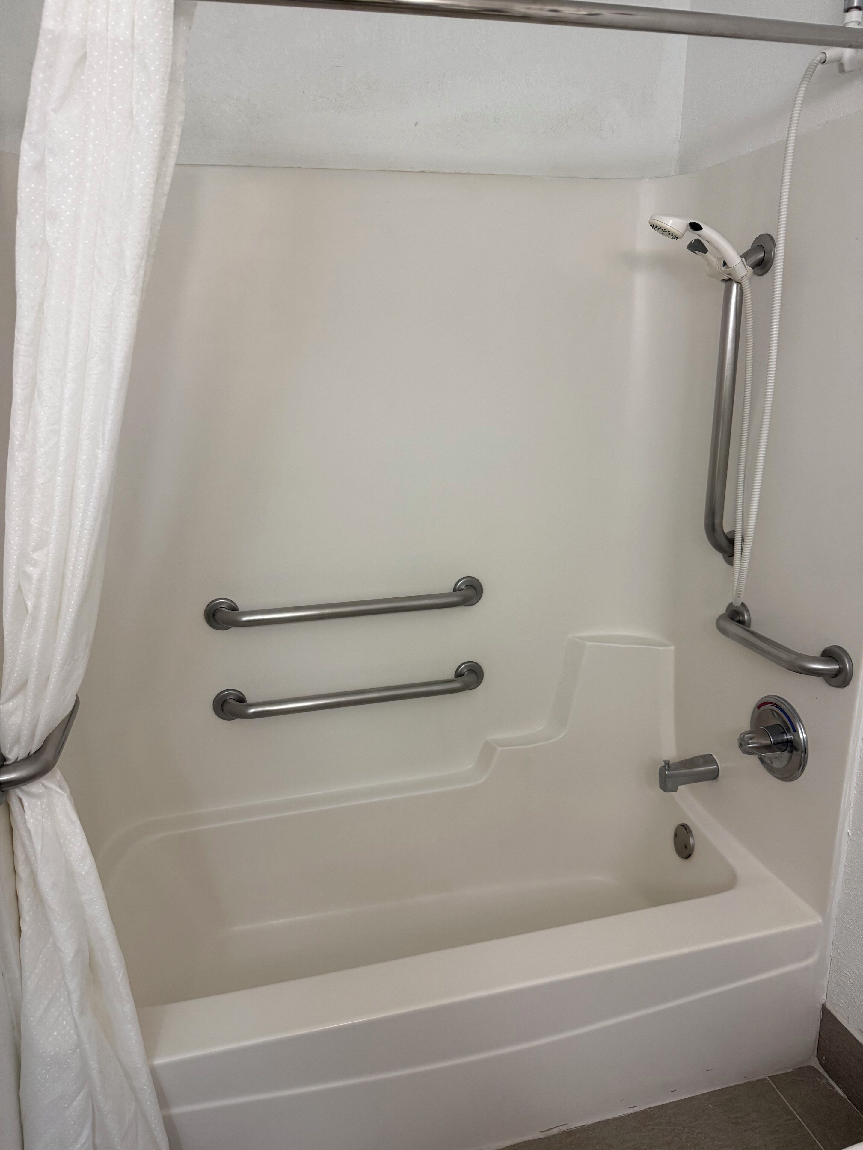 Una bañera blanca con barras de apoyo, cabezal de ducha y ducha de mano.