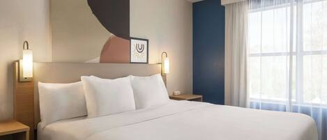 A neatly made bed with white linens, two bedside lamps, a wall art piece, and a large window with sheer curtains.