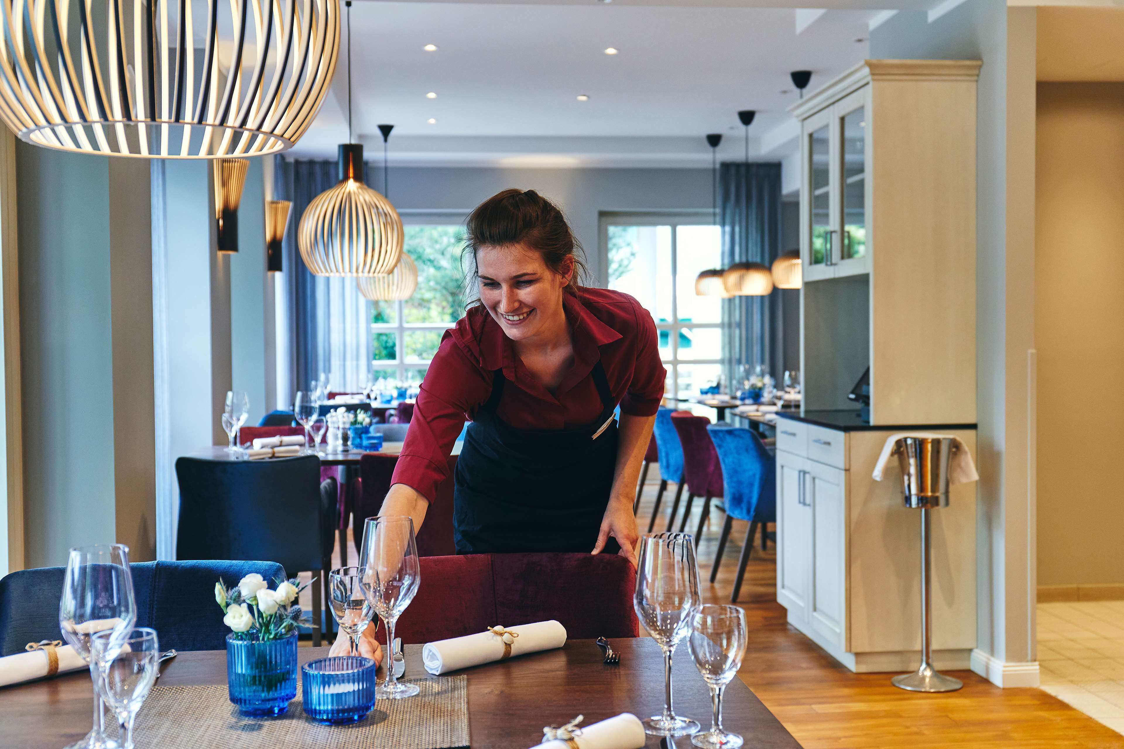 Une salle à manger d’hôtel avec une table dressée pour un repas, une personne qui prépare la table, et un décor intérieur moderne.