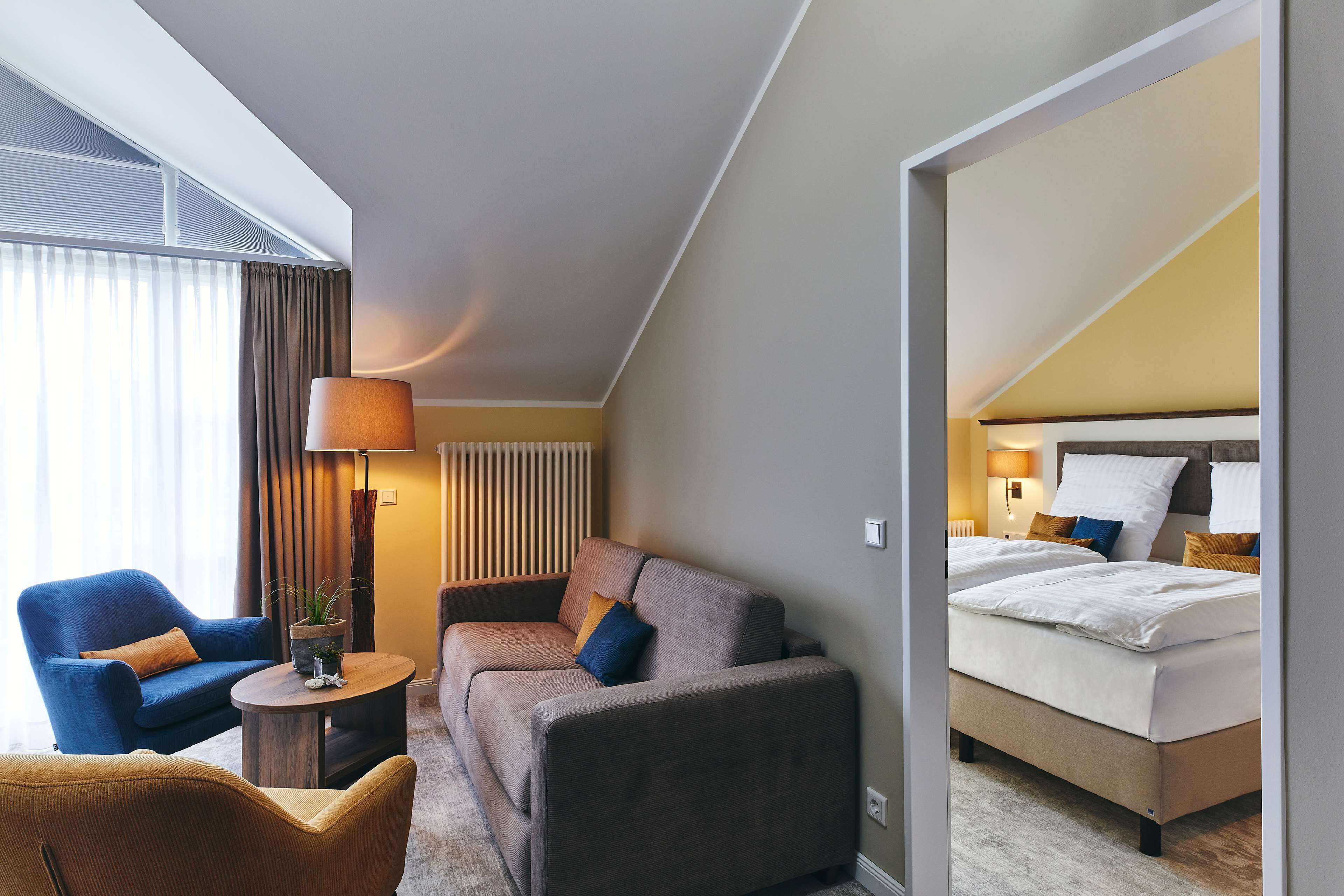 Une chambre d’hôtel avec un canapé, un fauteuil et un lit. On y trouve une lampe et une petite table sur laquelle est posée une plante.