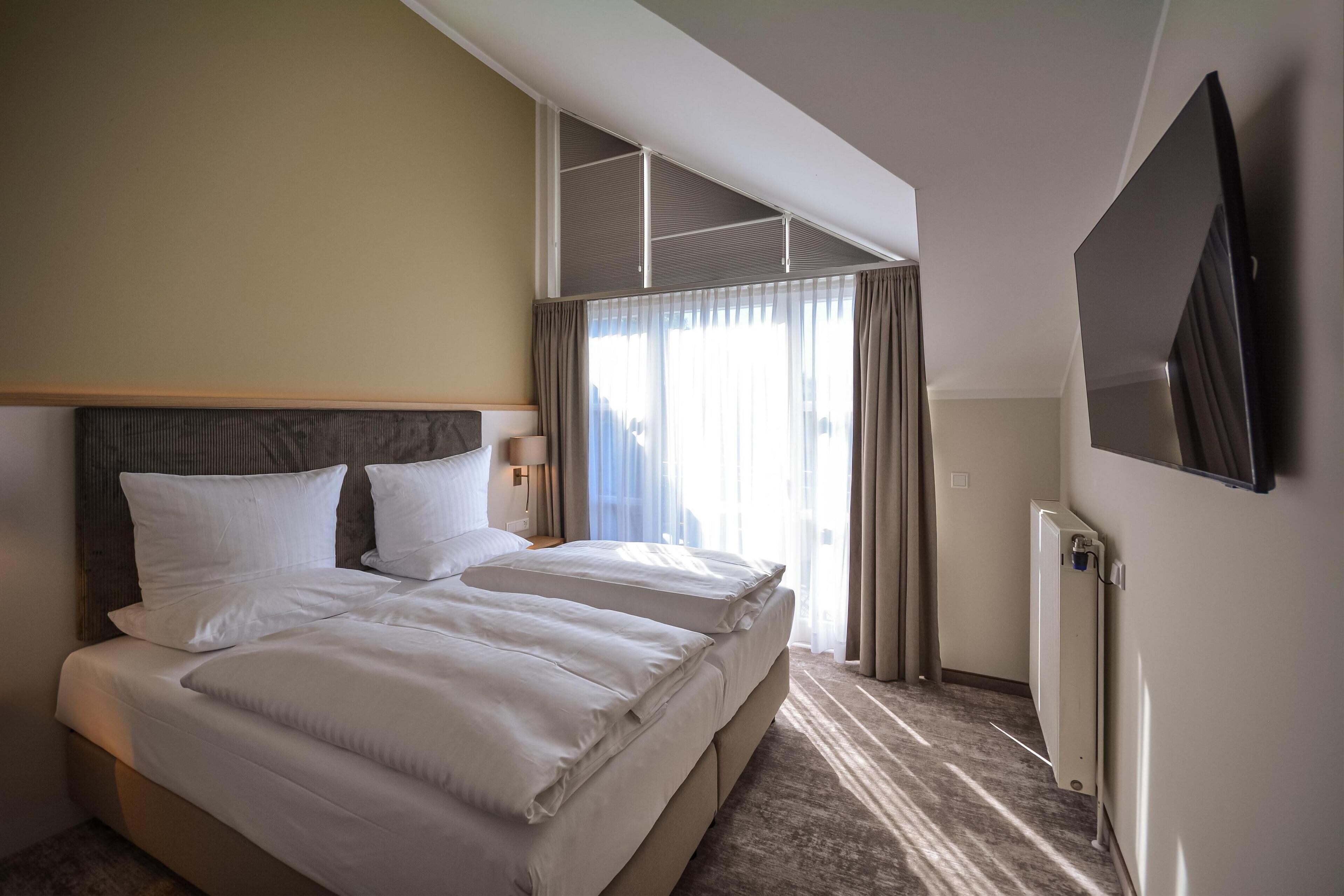 Ein Hotelzimmer mit einem großen Bett, einem an der Wand montierten Fernseher und einem Fenster mit Vorhängen.