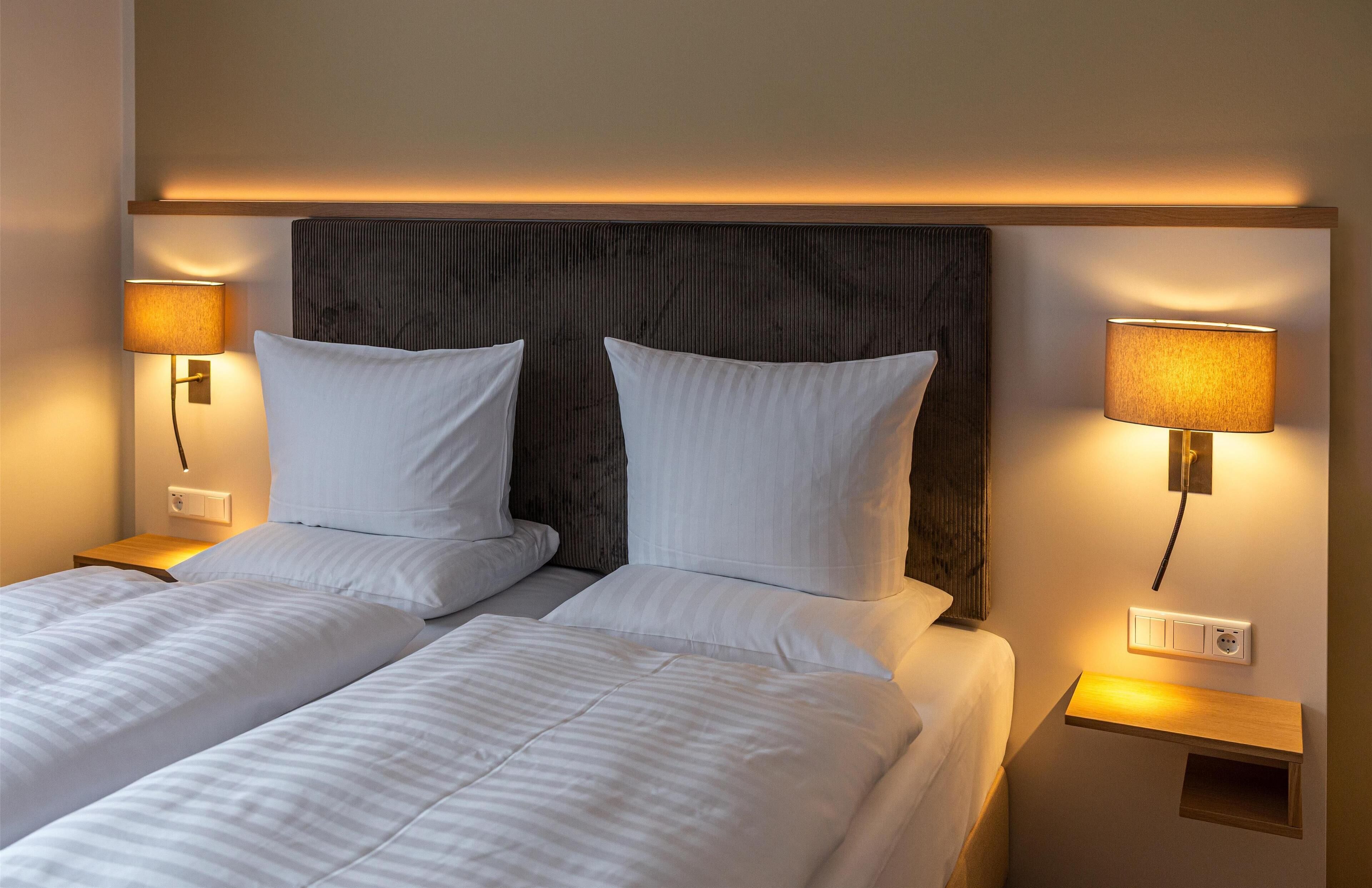 Une chambre d’hôtel avec deux lits, chacun agrémenté de draps et d’oreillers blancs, et flanquée de lampes fixées au mur.