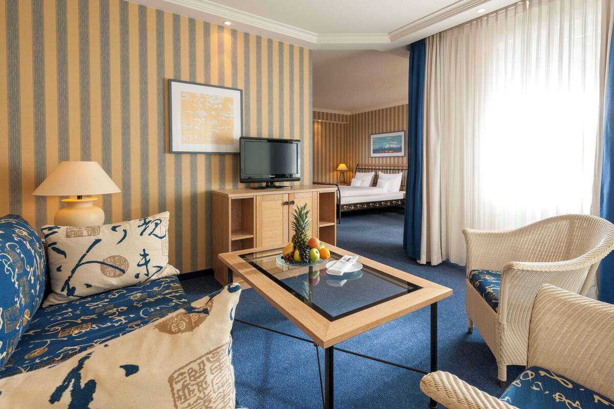 Ein Hotelzimmer mit gestreifter Tapete, einem Bett, einem Fernseher, einem Couchtisch mit Obst und einem Korbstuhl.