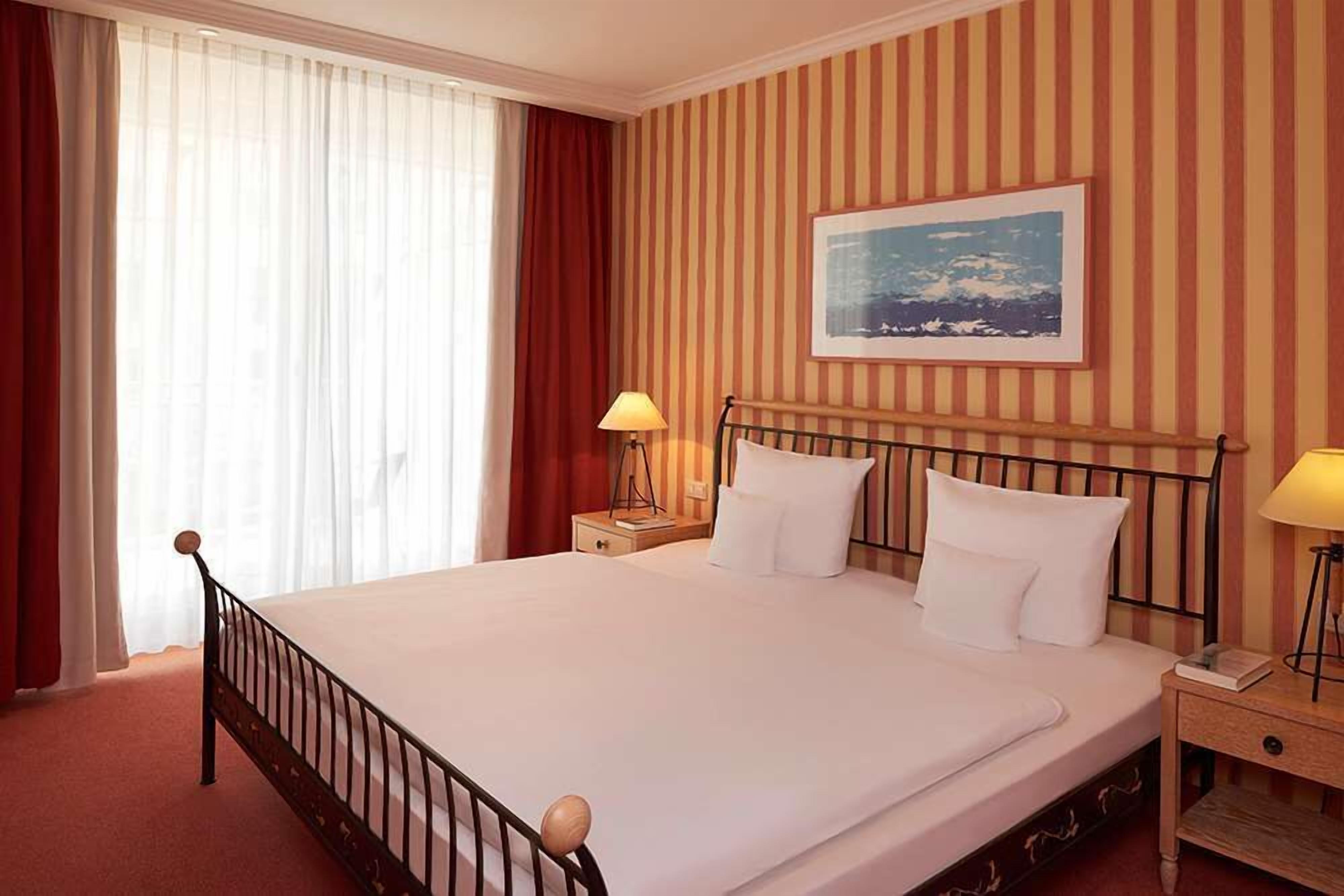 A hotel room with a large bed, two bedside tables, a nightstand, a window with curtains, and a striped wall.
