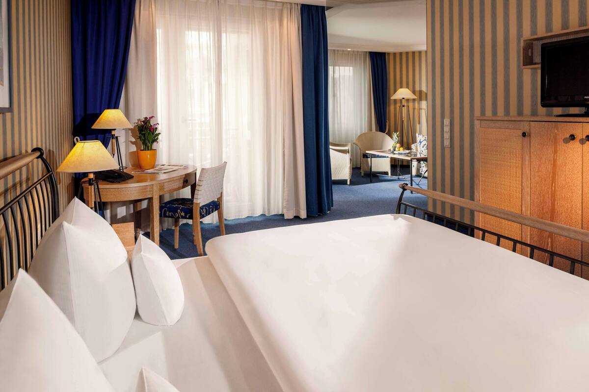 Ein Hotelzimmer mit einem großen Bett, einem Schreibtisch mit Lampe, einem Sessel und einem Fernseher auf einer Holzkommode.